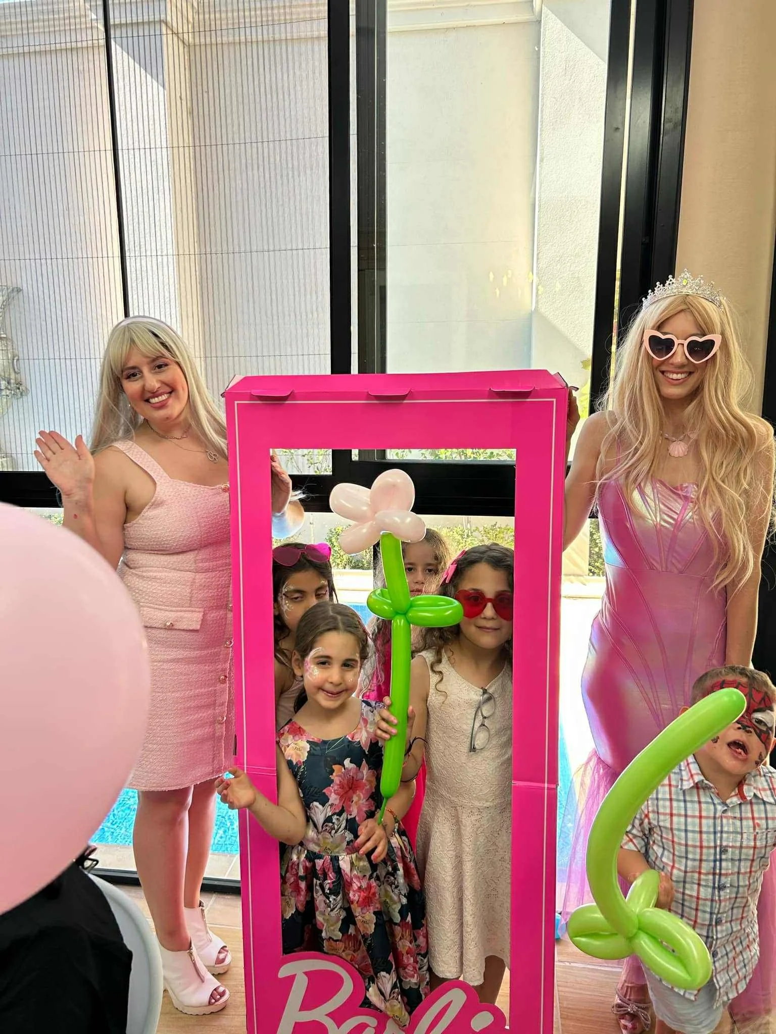 Pink castle-themed photo booth with children and two women in pink dresses, some children holding balloon flowers, inside a room with large windows, one woman wearing heart-shaped glasses and tiara, children with face paint and colorful outfits.