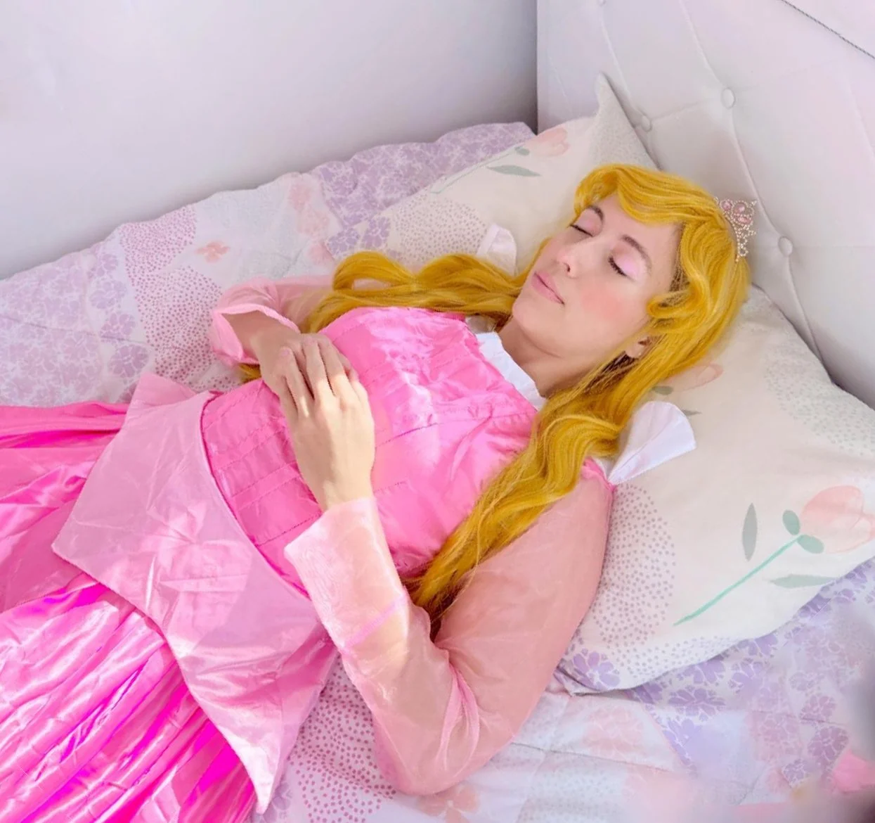 A woman dressed as Aurora Sleeping Beauty, with long blonde hair, wearing a pink princess dress and a tiara, sleeping peacefully on a bed with floral-patterned sheets.