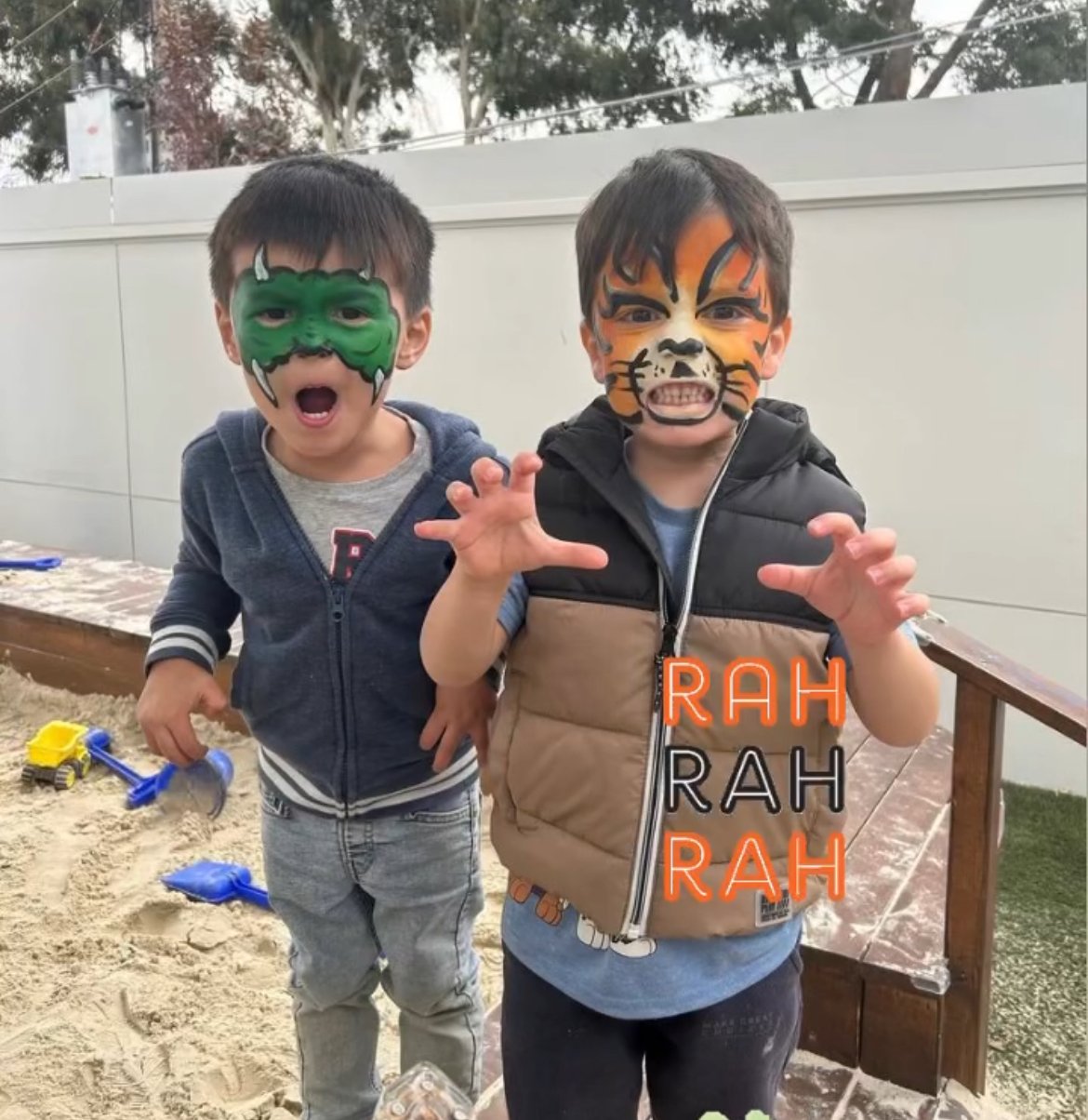 Two young boys with face paint in a playful outdoor setting. The boy on the left has a green Hulk face paint, and the boy on the right has a tiger face paint. They are making clawing gestures and expressions of fierce playfulness, with sand and toy shovels on the ground behind them.