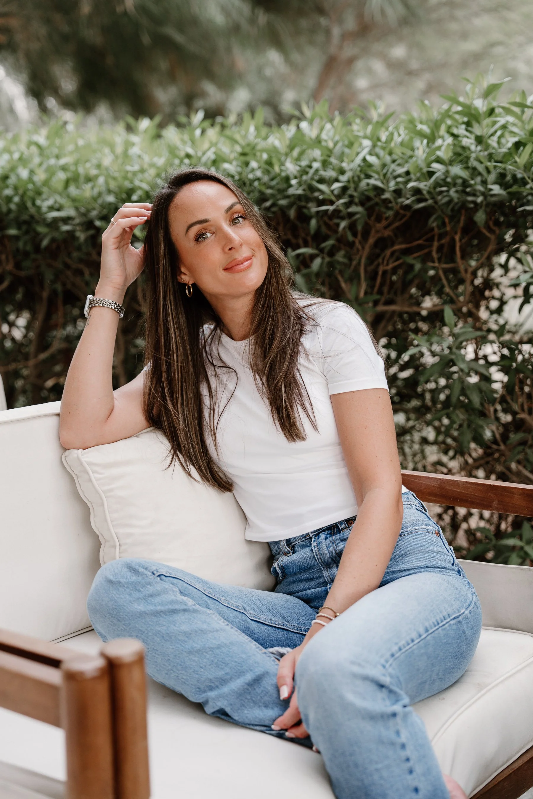 Young woman sitting on outdoor couch with green bushes in the background, wearing a white t-shirt and jeans.