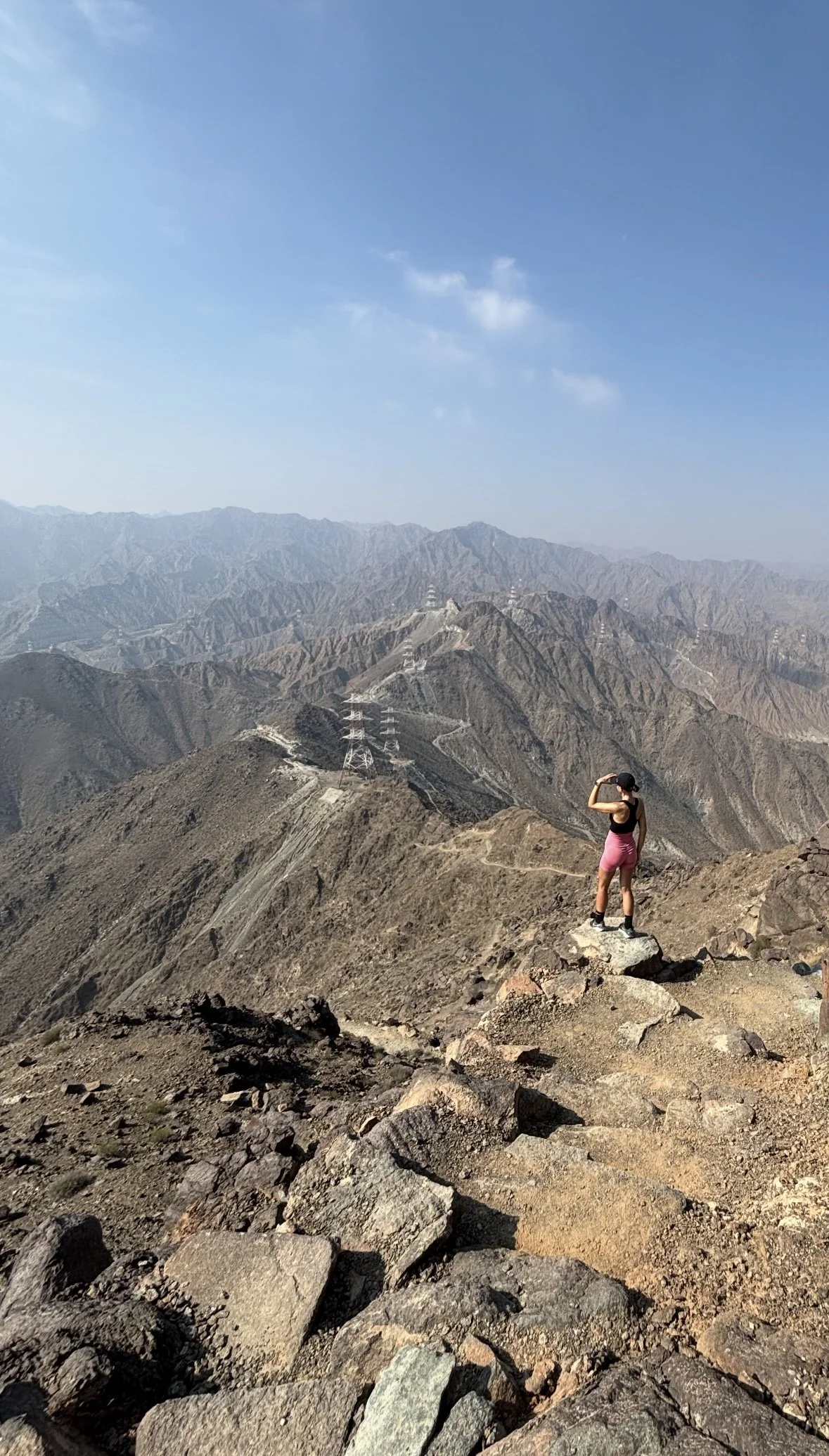 A woman standing on a rocky mountain ridge, looking out over a vast range of mountains under a clear blue sky.