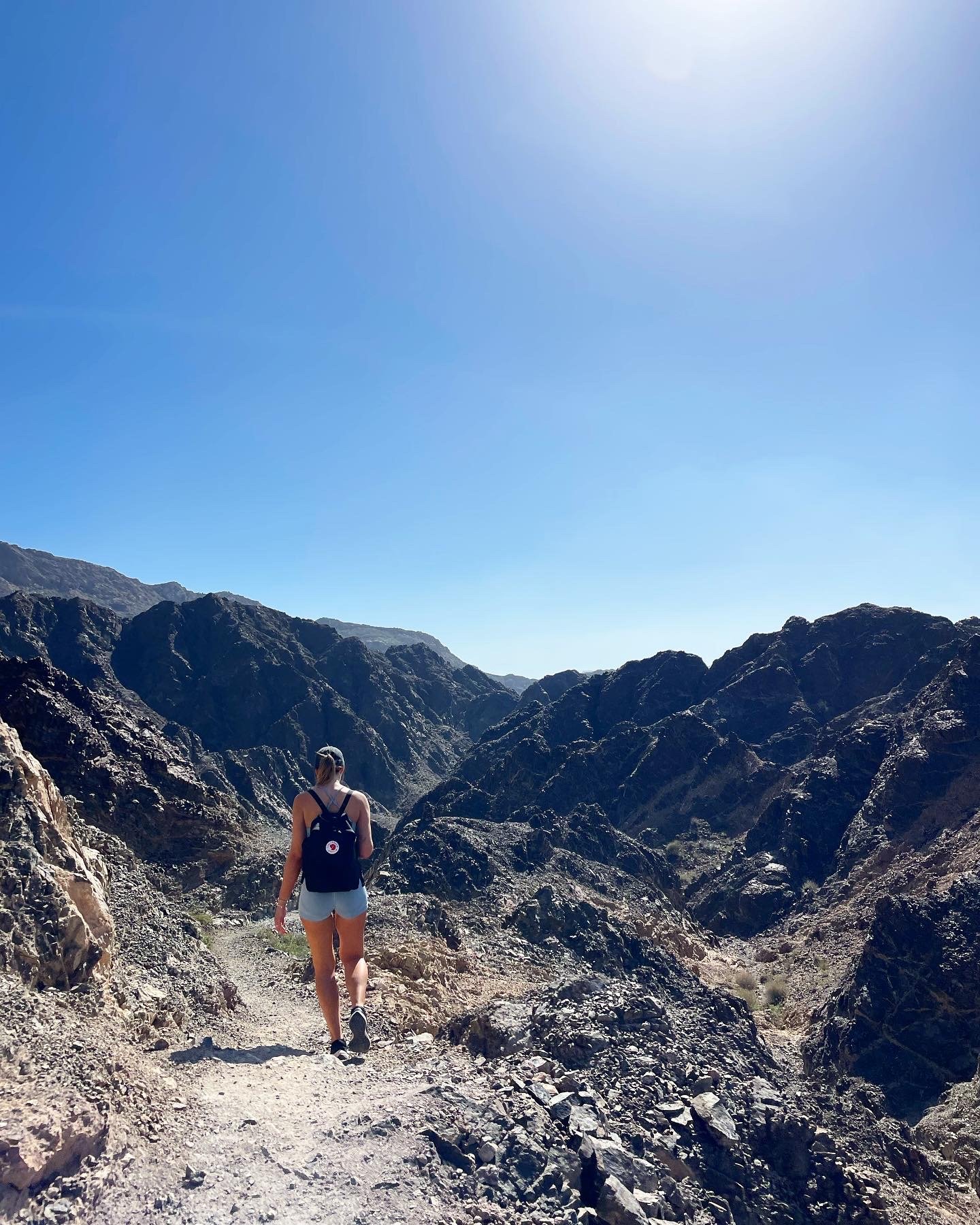 A woman hiking on a rocky trail in a mountainous desert landscape under a clear blue sky.