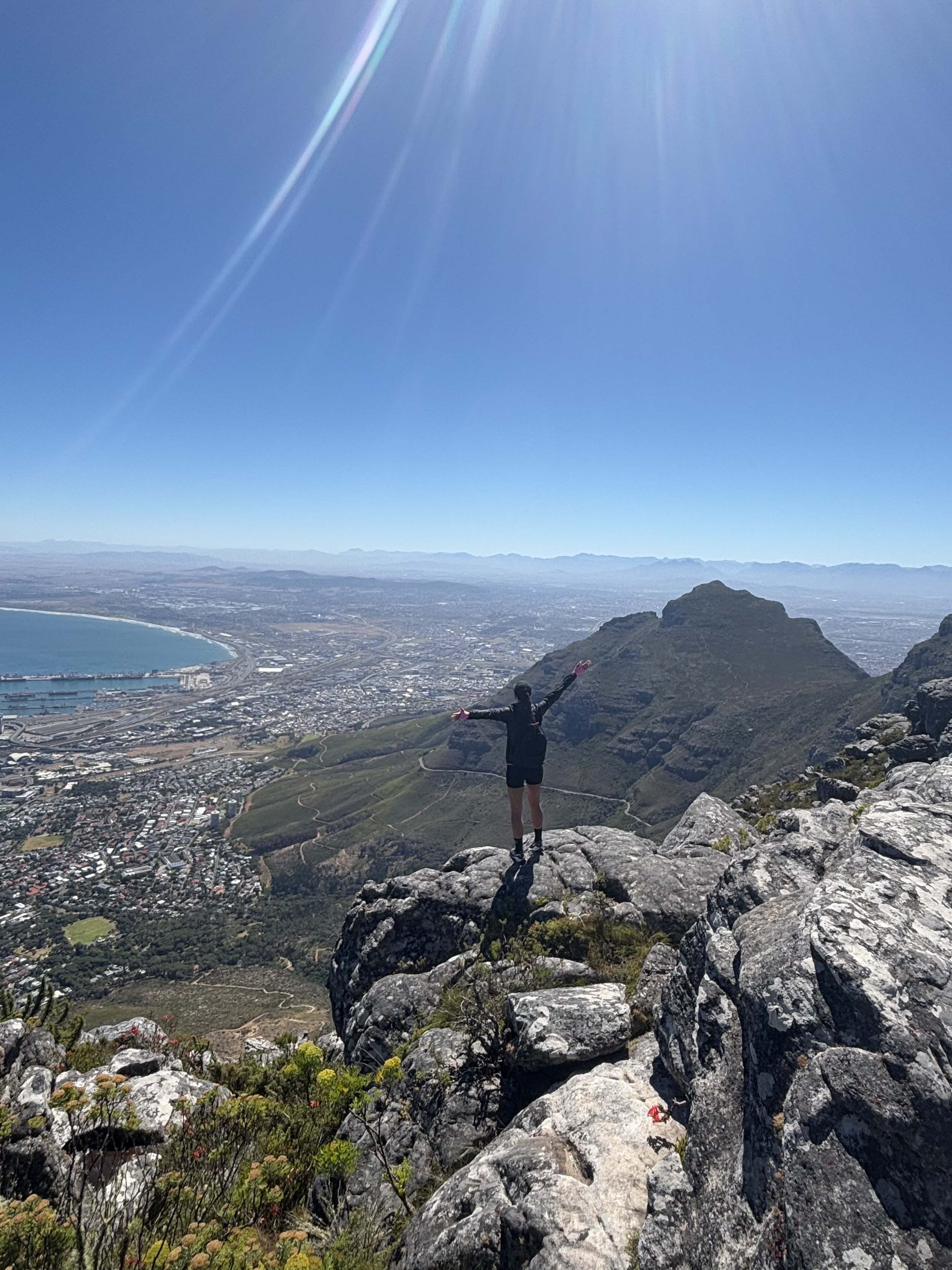 Person standing on rocky mountain peak with arms outstretched, overlooking a city and ocean on a sunny day.