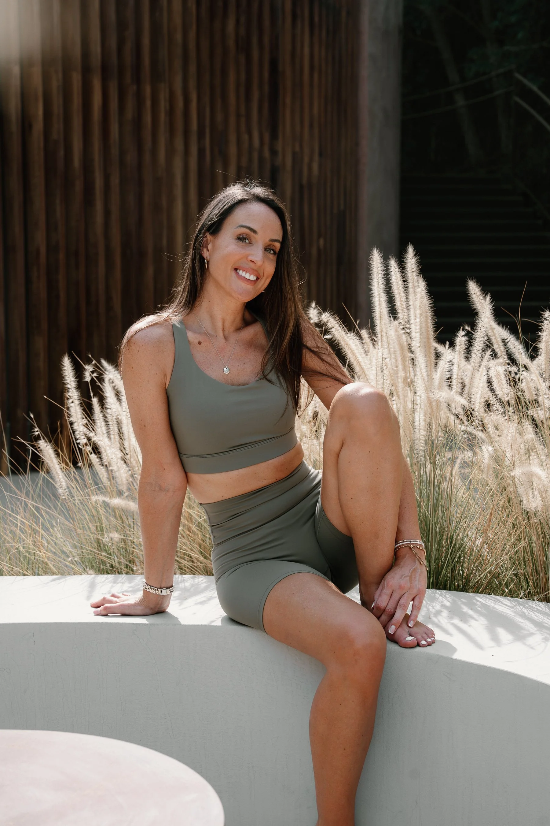 A woman in a gray sports bra and shorts sitting outdoors on a white bench, smiling with her legs crossed, surrounded by tall, white feathery grasses.