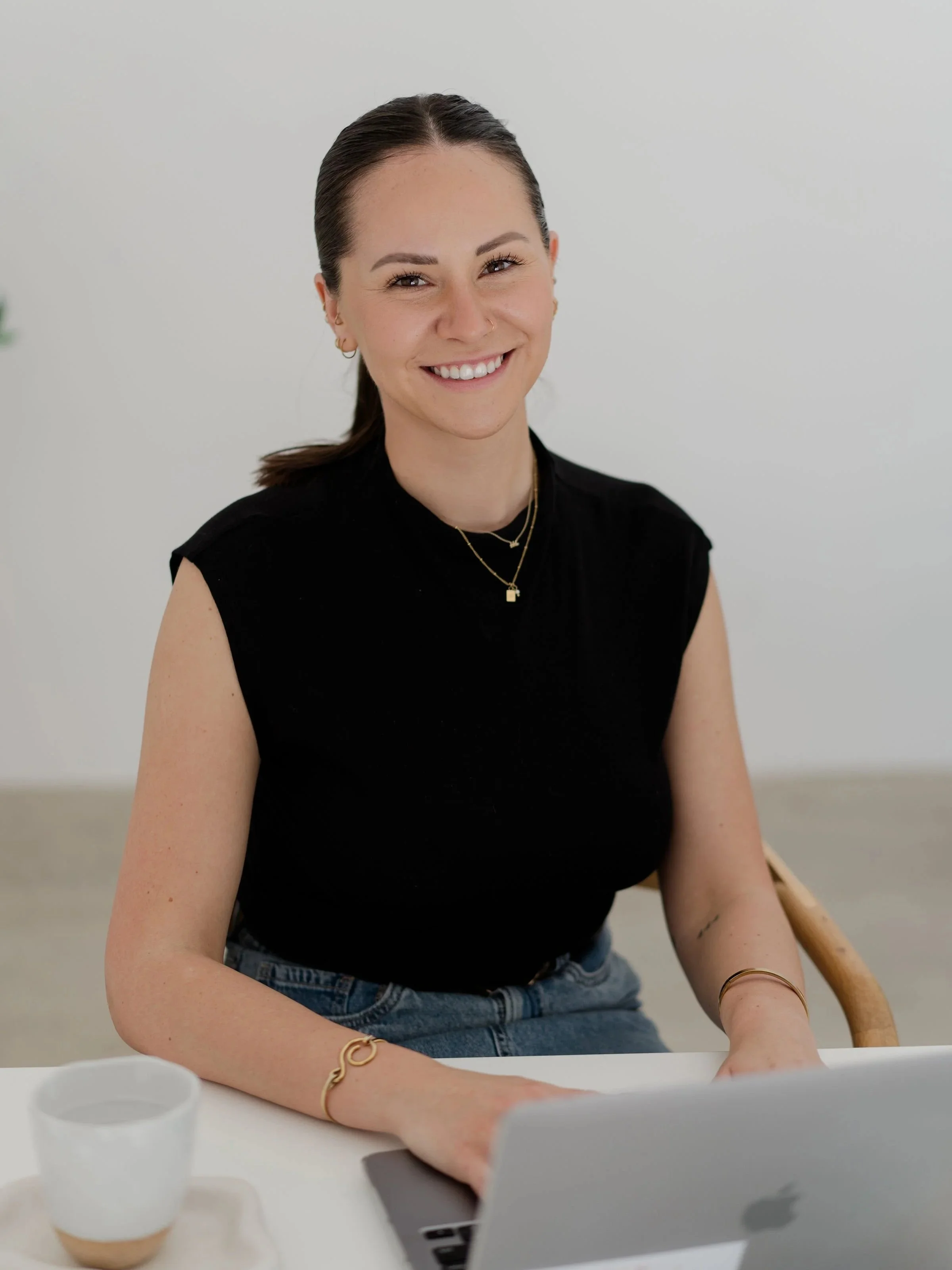 A woman sitting at a table with a laptop and a coffee cup, smiling at the camera.