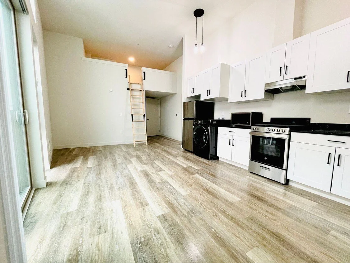 Modern kitchen with white cabinets, black hardware, stainless steel appliances, and light wood flooring, featuring a loft area with a wooden ladder.