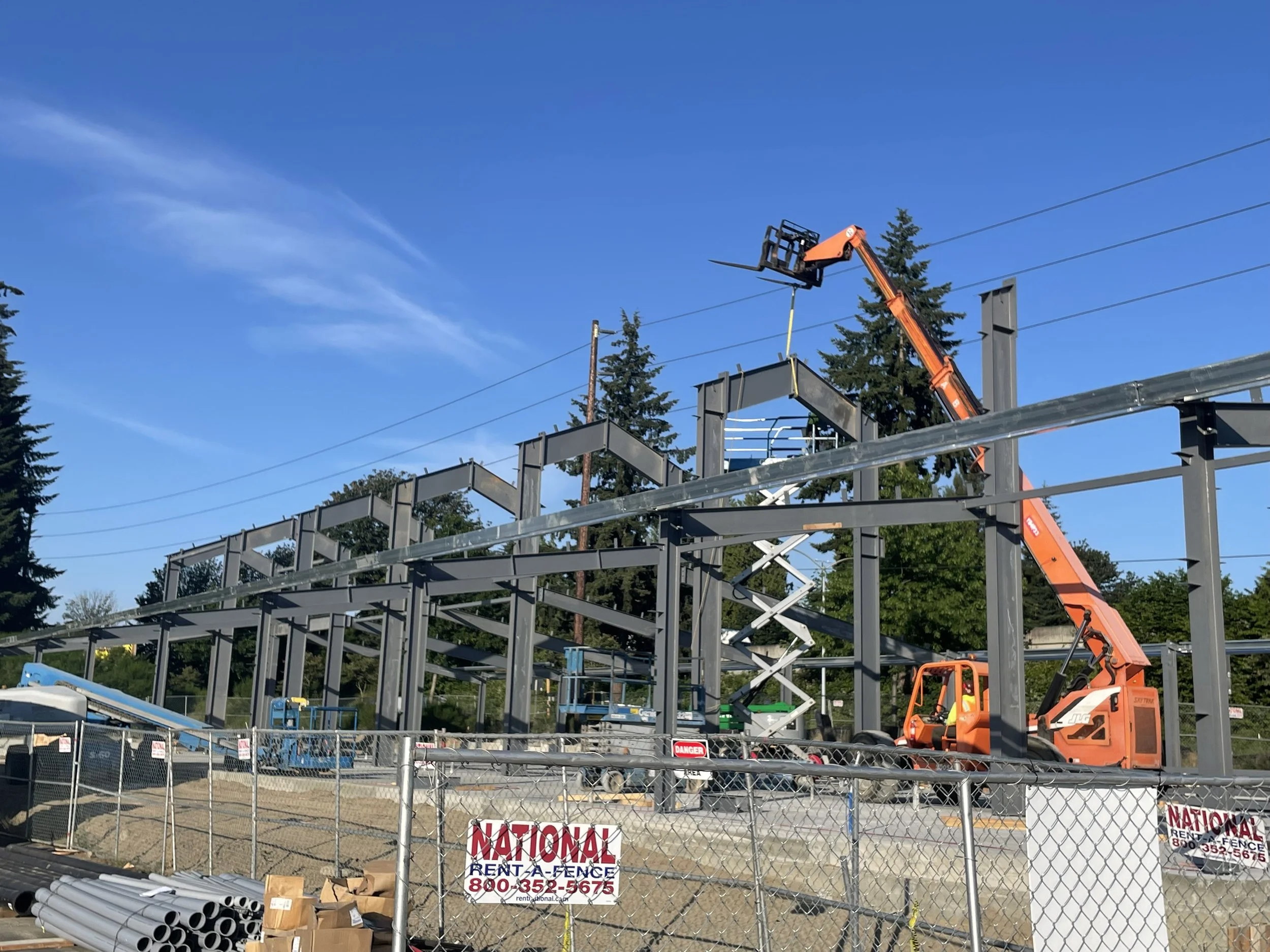 Construction site with steel framework being assembled, orange crane lifting steel beams, and a chain-link fence in the foreground with signs for a fencing company.