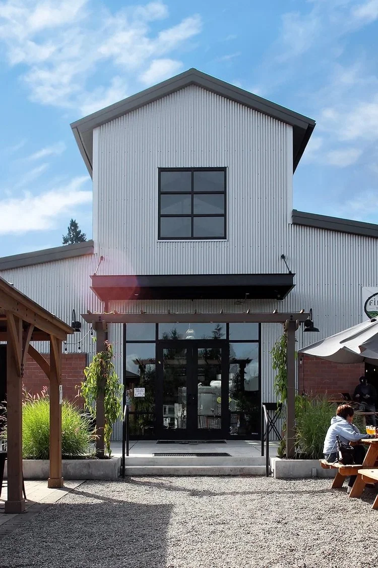 Exterior of a modern two-story building with corrugated metal siding, large glass doors, and a black-framed window on the upper level. There are outdoor seating areas with a person sitting at a picnic table.