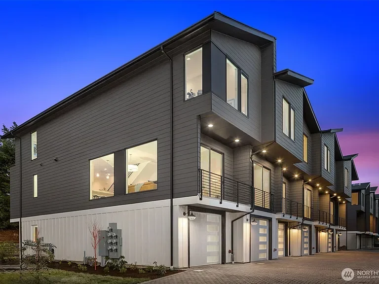 Modern kitchen with light wood cabinets, stainless steel appliances including a dishwasher, oven, and refrigerator, and a laundry area with a washing machine and dryer in the background. The room has wood-look flooring and white walls with small windows.