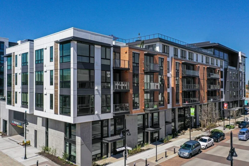 Modern kitchen with light wood cabinets, stainless steel appliances including a dishwasher, oven, and refrigerator, and a laundry area with a washing machine and dryer in the background. The room has wood-look flooring and white walls with small windows.