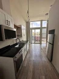 A modern kitchen with white cabinets, a microwave, a stove, a black countertop, and a refrigerator, leading to a balcony with a view of the outdoors.