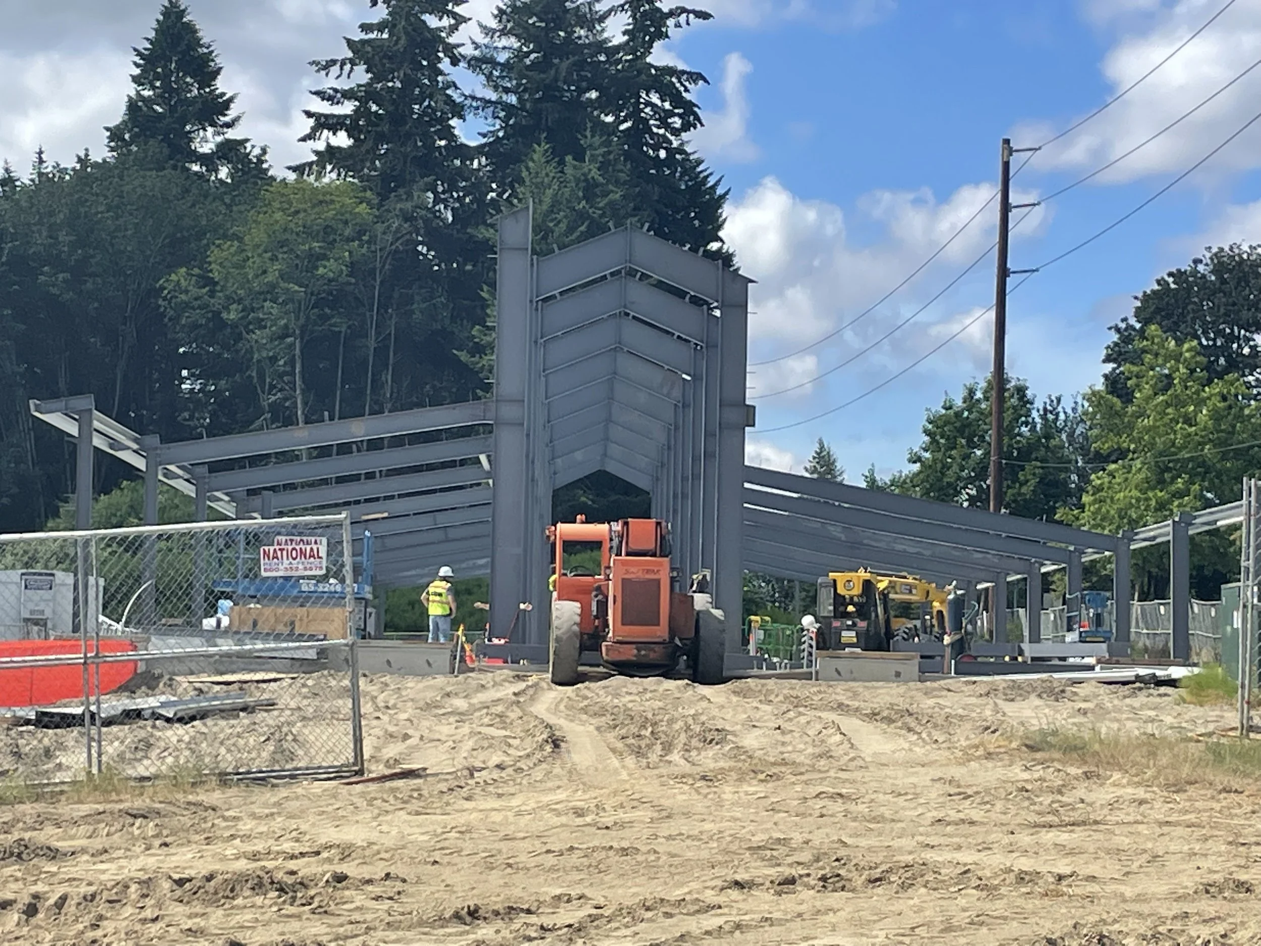 Construction site with a steel structure frame being assembled, construction workers, and heavy machinery, set against a backdrop of trees and a partly cloudy sky.