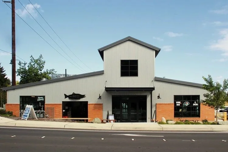 A modern building with a metal siding, brick accents, and large windows, featuring a fish decoration on the left window and a sign indicating it is a wine bar and shop, located along a street with visible utility poles and trees.
