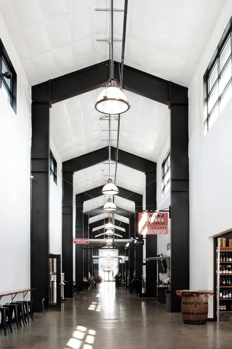 Long indoor hallway with high, arched ceiling and large black support beams, white walls, industrial lighting fixtures hanging from the ceiling, and windows letting in natural light.