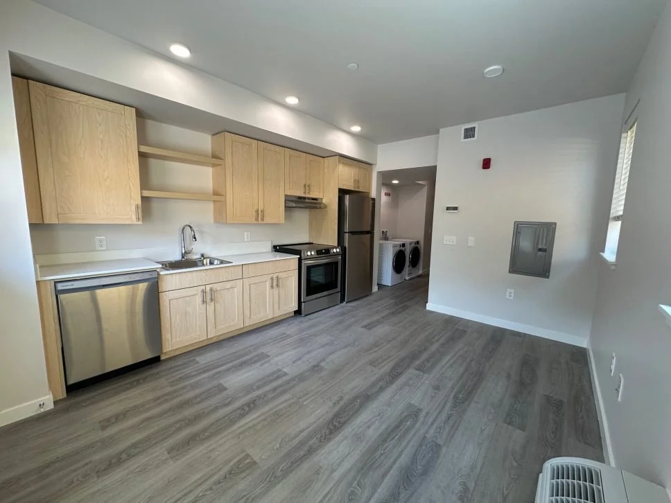 Modern kitchen with light wood cabinets, stainless steel appliances including a dishwasher, oven, and refrigerator, and a laundry area with a washing machine and dryer in the background. The room has wood-look flooring and white walls with small windows.