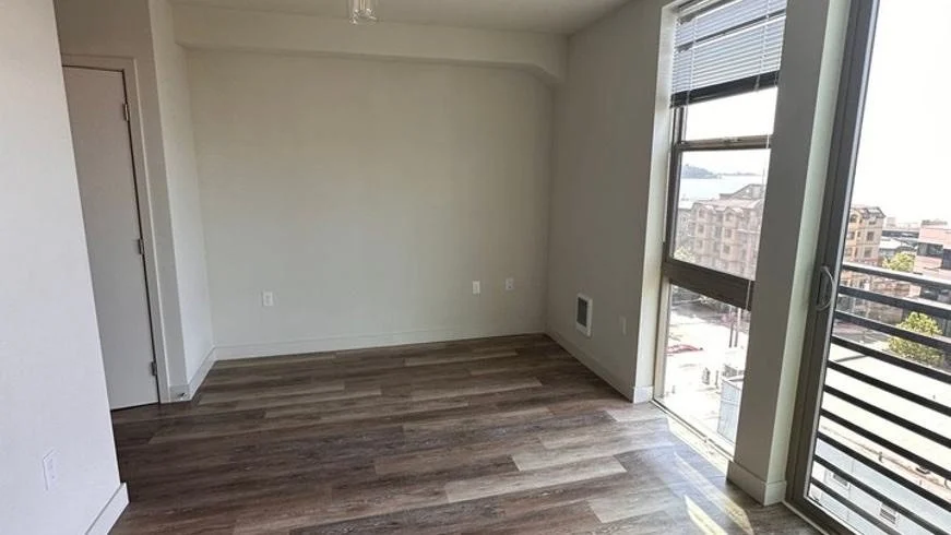 Completed room with hardwood floors, white walls, large windows with blinds, and a sliding glass door leading to a balcony.