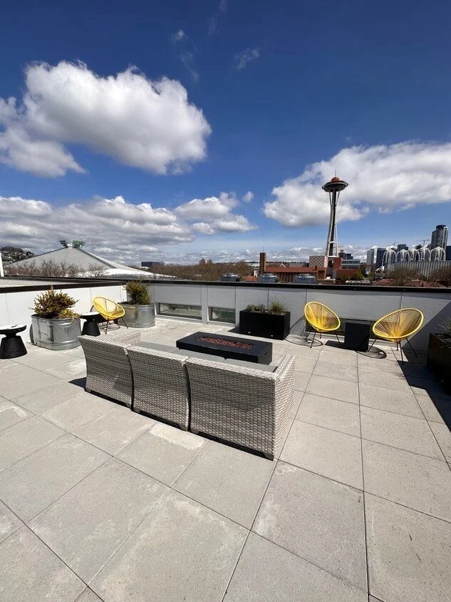 Rooftop patio with patio furniture, yellow chairs, black planters, and city skyline with the Space Needle in Seattle under a partly cloudy sky.