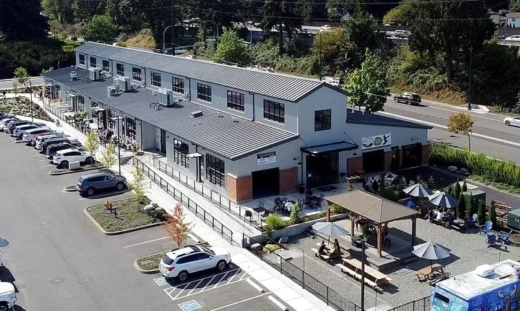Commercial building with outdoor seating and parking lot, surrounded by trees.