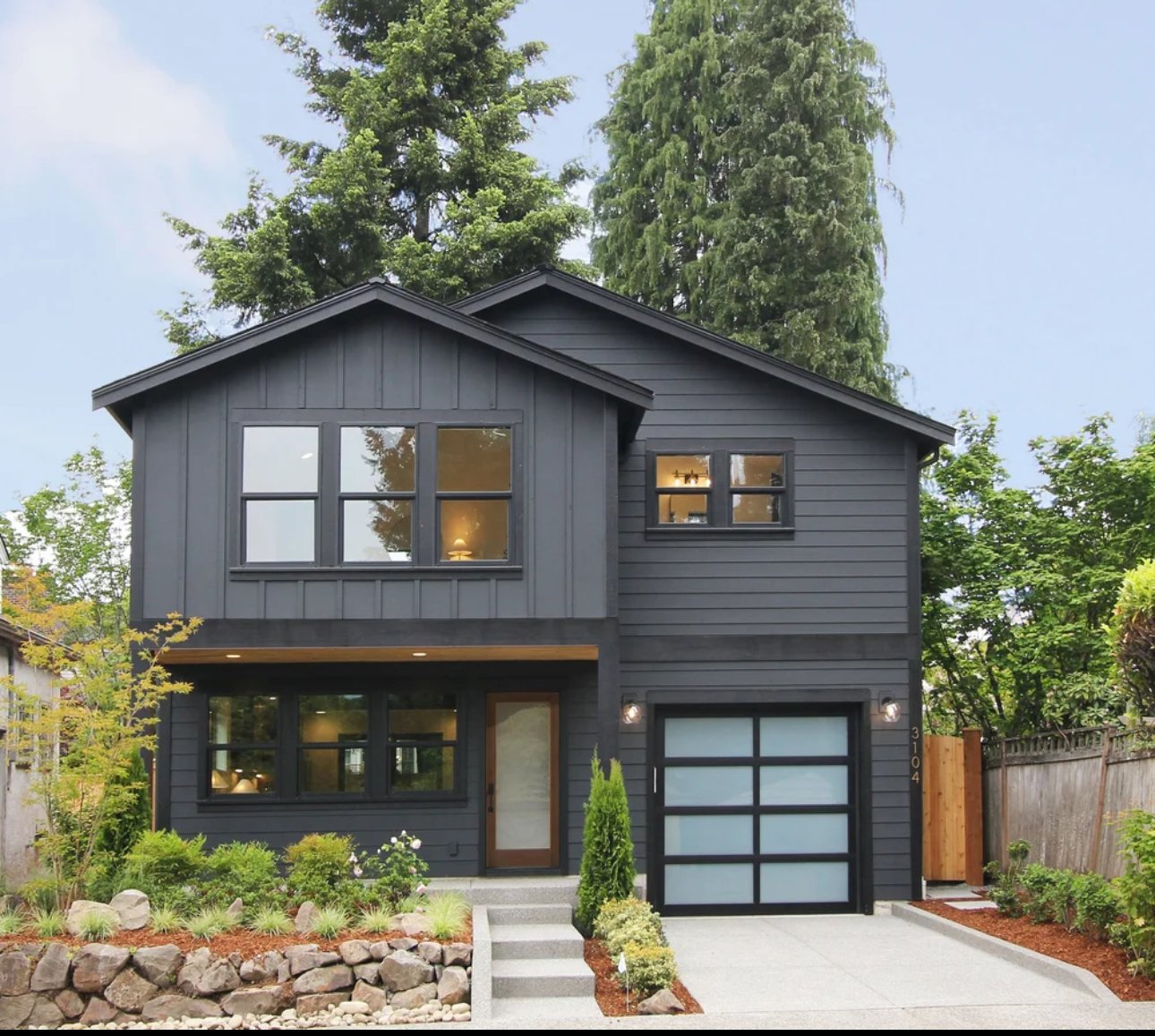Front view of a modern two-story house painted dark gray with large windows, a garage, and a landscaped front yard with small bushes, flowers, rocks, and a concrete walkway.