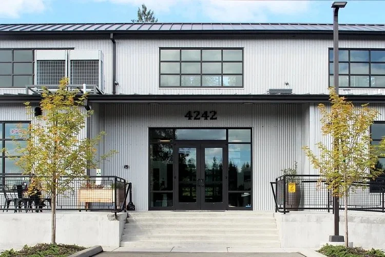 Modern industrial-style building with a gray metal exterior, numbered 4242, and glass doors at the entrance, flanked by two trees and a small ramp for accessibility.