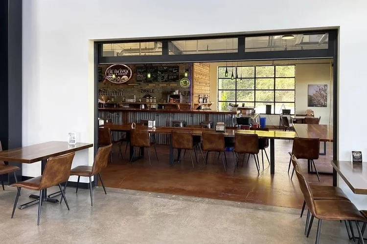 Coffee shop with brown chairs and tables, a counter with a menu and coffee equipment, and a large window with natural light.