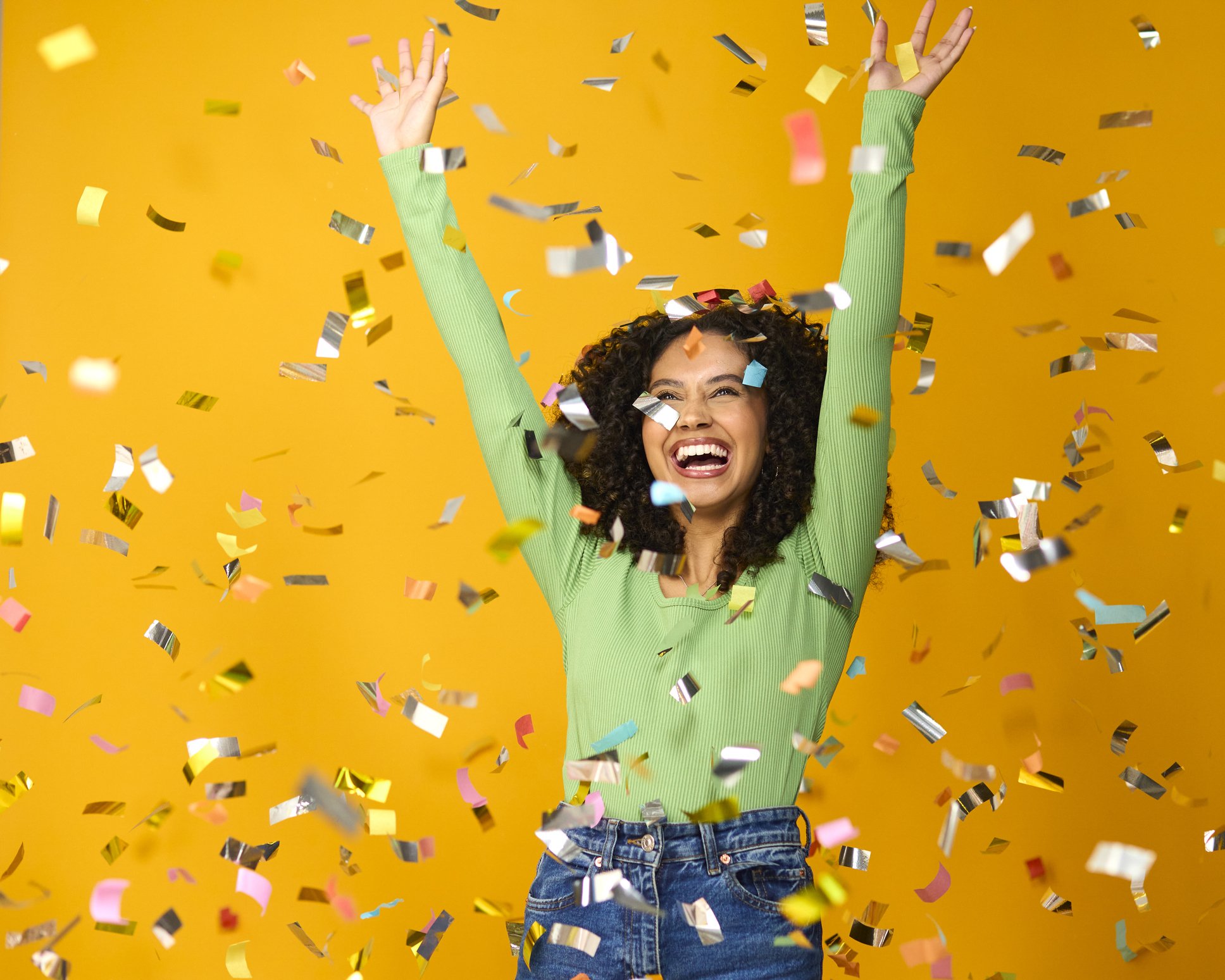 A woman with curly dark hair smiling and raising her arms in celebration, surrounded by gold, silver, pink, and blue confetti against a bright yellow background.