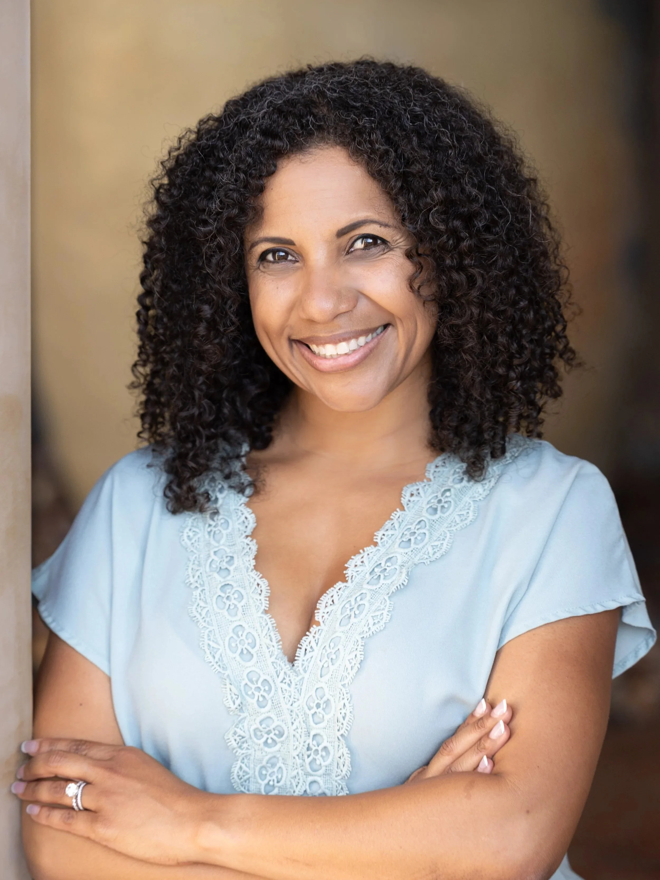 A woman with dark, curly hair smiling, wearing a blue top with lace details, arms crossed, standing in front of a beige background.