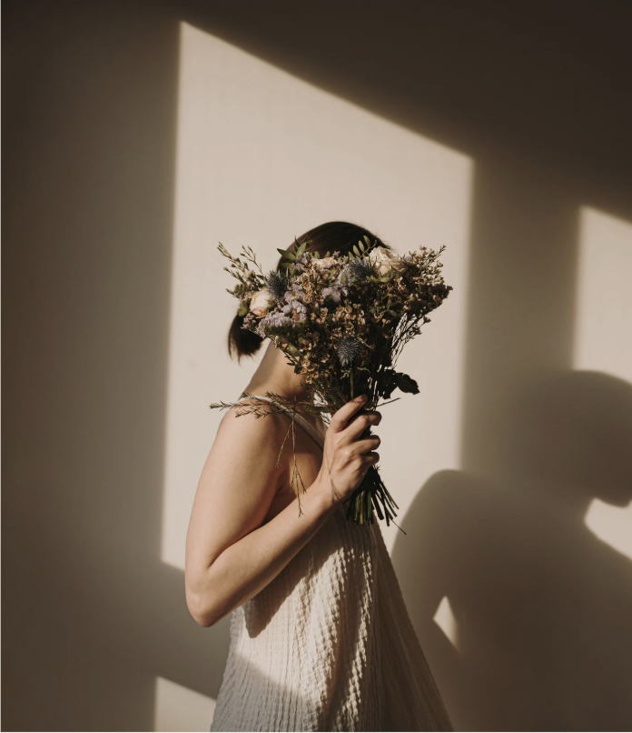 Une personne tient un bouquet de fleurs devant le visage, dans une pièce éclairée par la lumière naturelle. The person porte une robe blanche, et l'installation lumineuse crée un contraste entre l'ombre et la lumière.