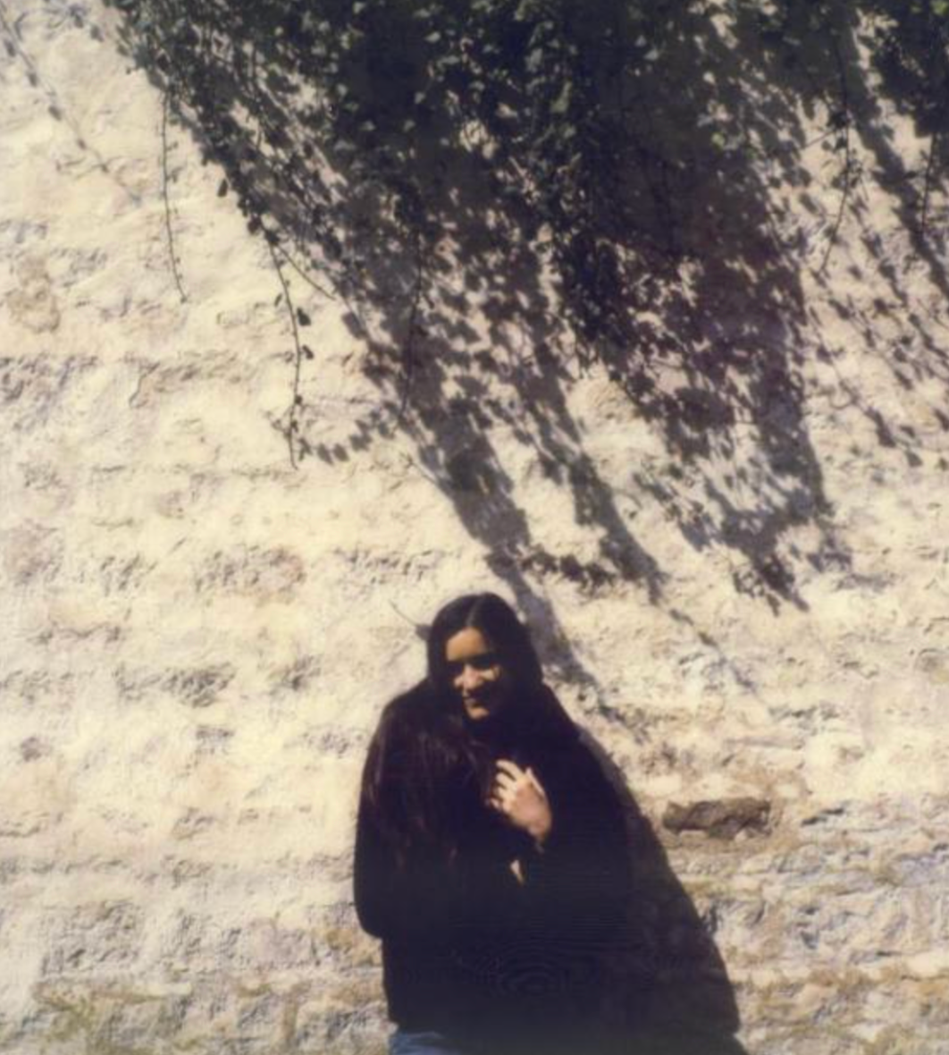 Une femme posant contre un mur en pierre avec un arbre projetant une ombre sur le mur.