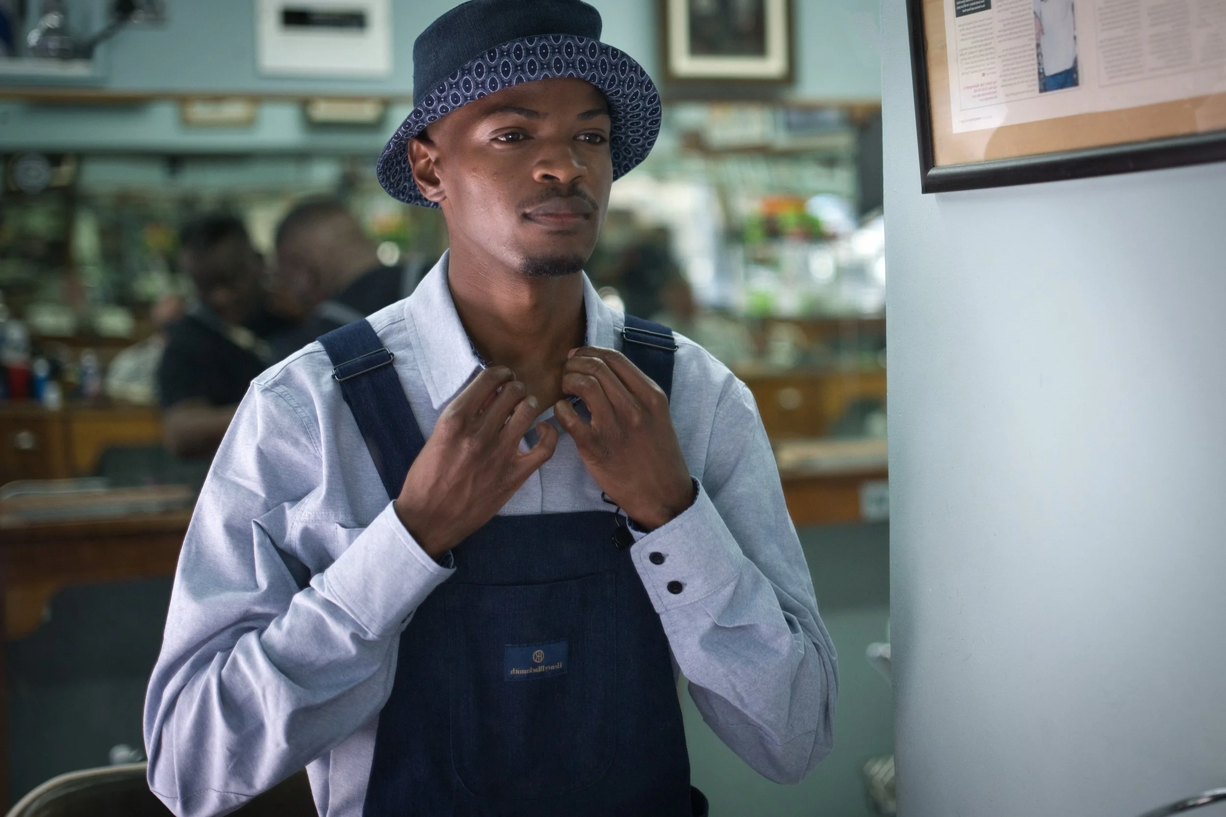 Henry Blacksmith clothing shoot. Brand development for clothing company. A young man adjusting his shirt collar, wearing a blue apron and a patterned bucket hat, standing inside a cafe or restaurant.