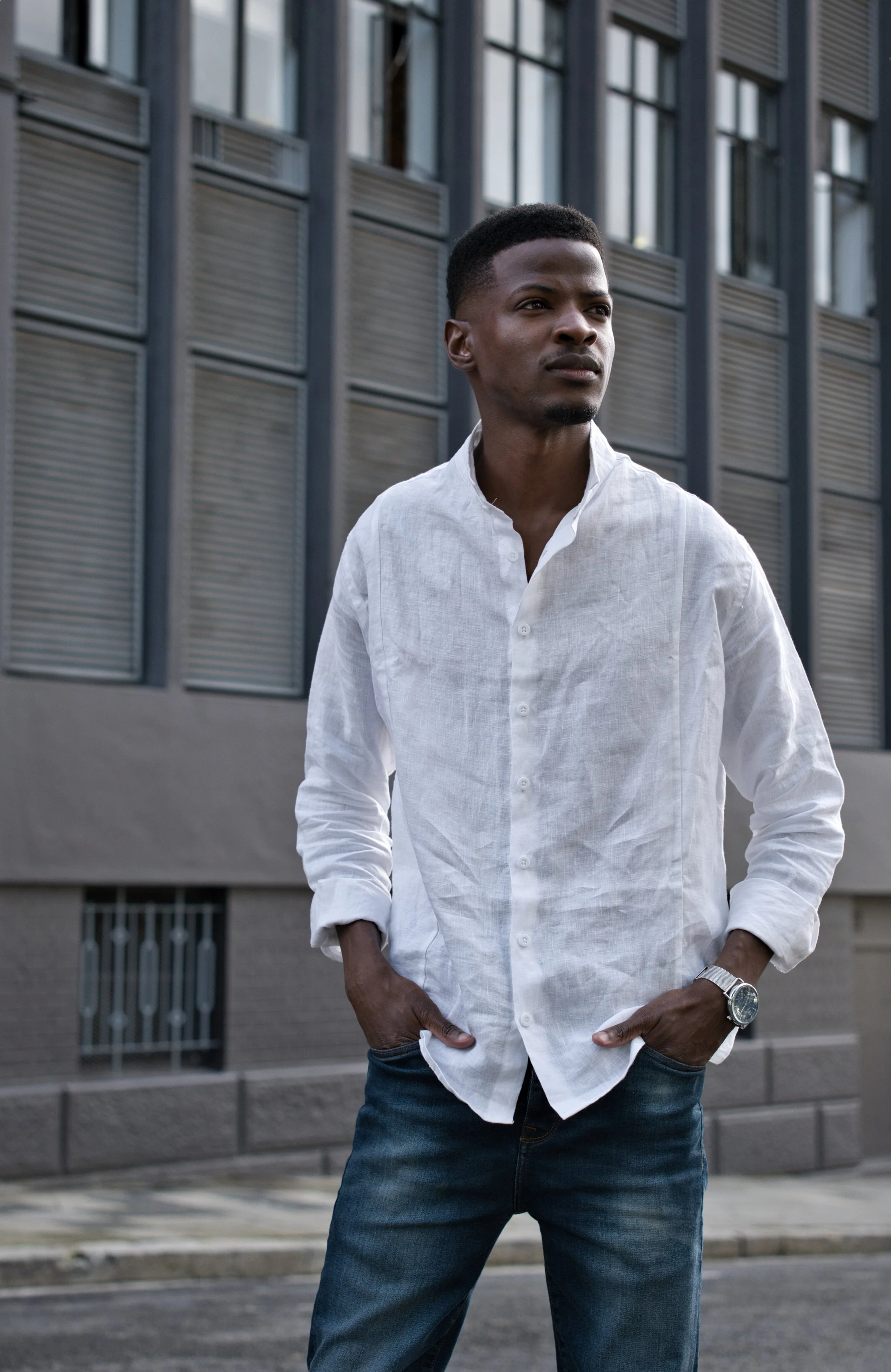 Henry Blacksmith product shoot. A man with dark skin, short black hair, and a goatee wearing a white button-up shirt with rolled-up sleeves and dark jeans stands outdoors.