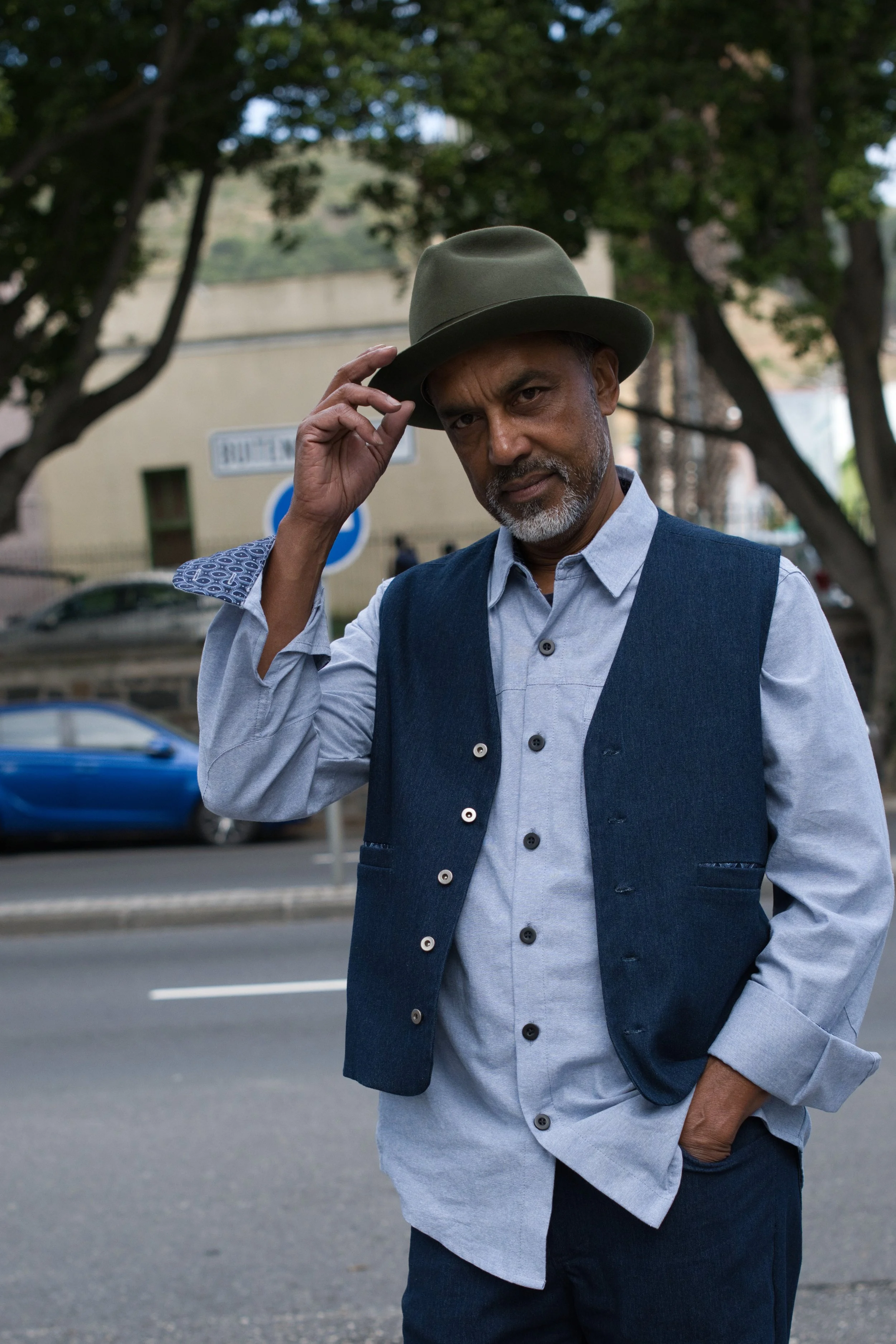 Henry Blacksmith product shoot. A middle-aged man with a salt-and-pepper beard, wearing a green fedora, a blue vest over a light blue shirt, and dark pants, standing outdoors