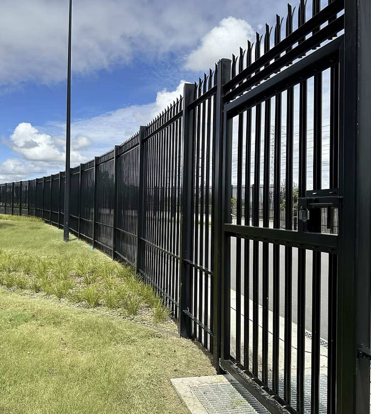 Black metal fence along a sidewalk with small bushes planted in the ground underneath, trees in the background, and a cloudy sky.