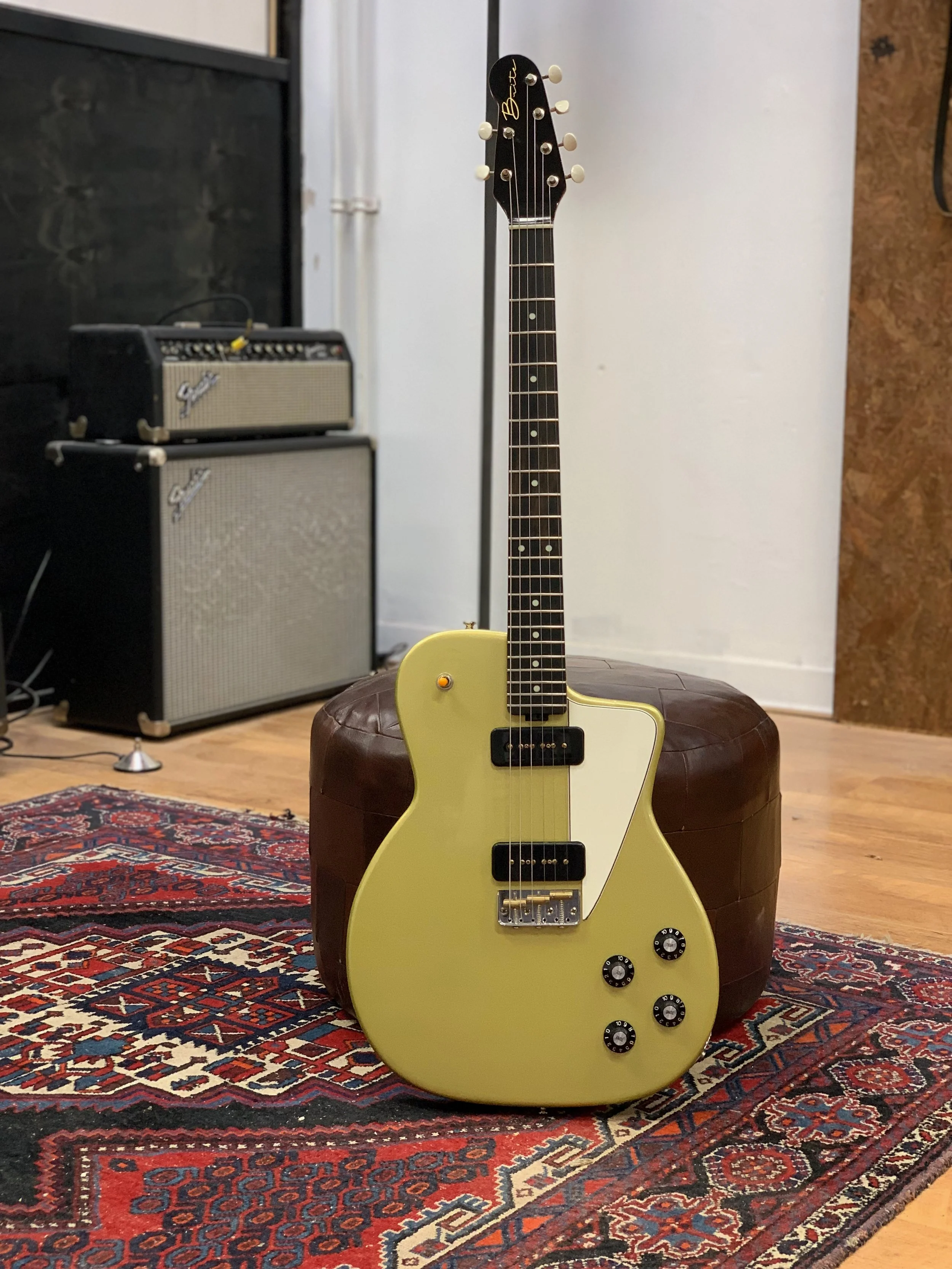 Een gele elektrische gitaar met zwarte knoppen staat op een ottomaanse poef, op een kleurrijk geborduurd tapijt in een kamer met houten vloer.