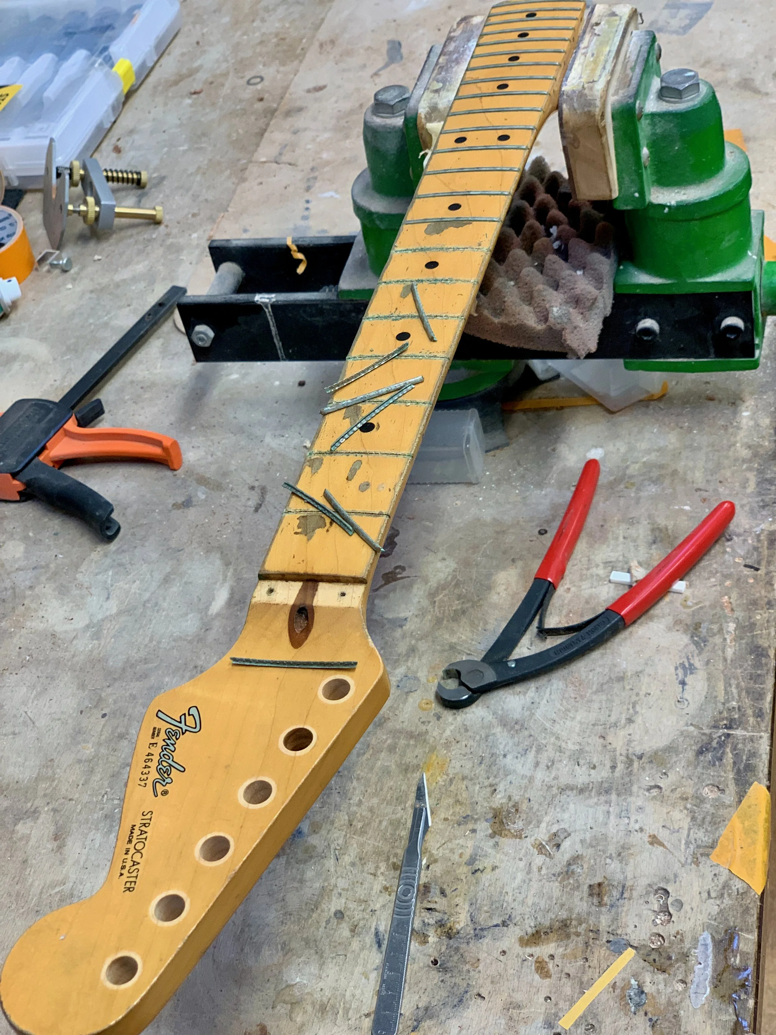 Een werkbank met een gitaarhals die op een steun geplaatste. Op de werkbank liggen gereedschap en kleine onderdelen, zoals ratels en vijlen, voor het repareren of bouwen van de gitaar.