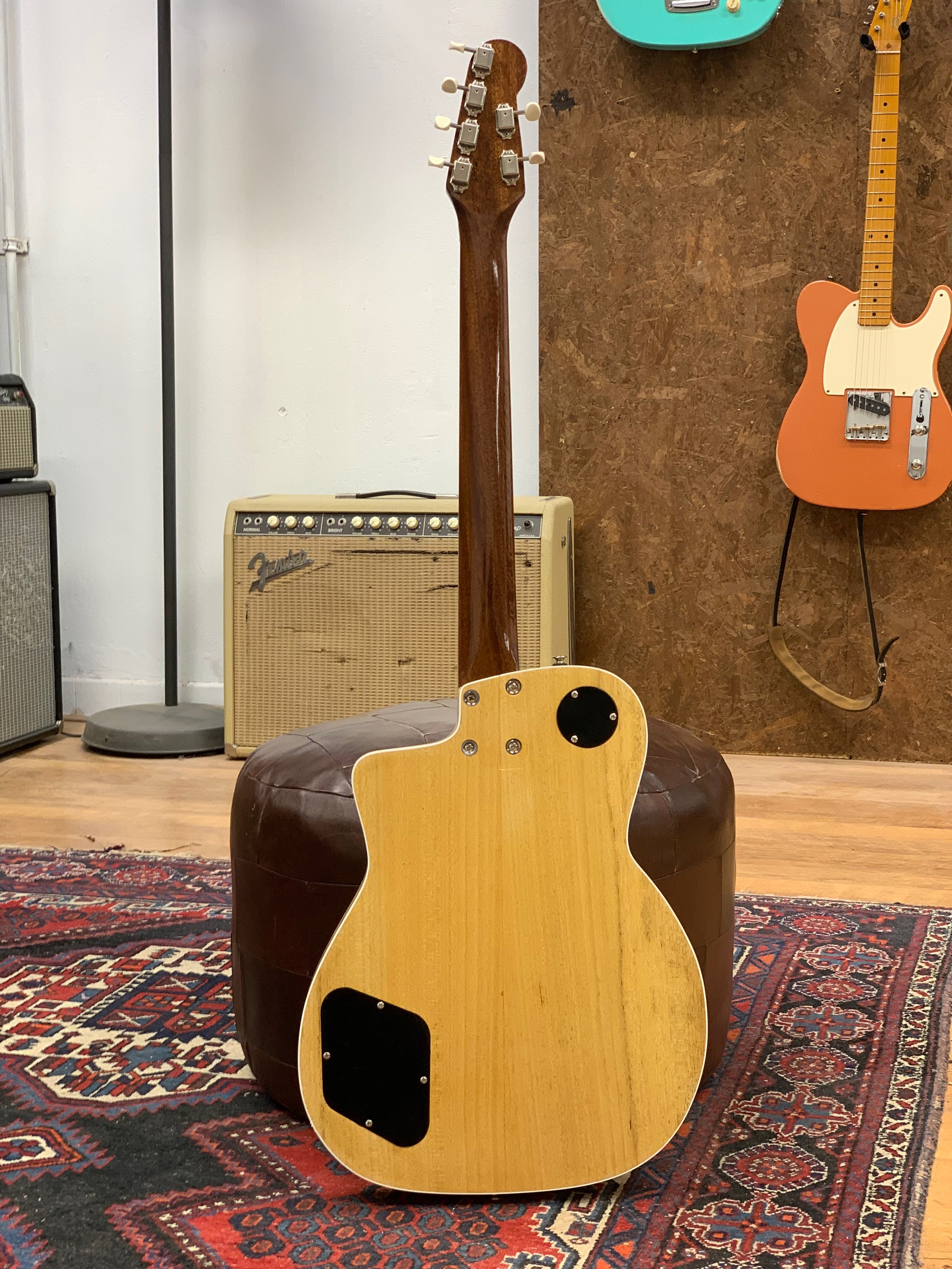 Binnenzijkant van een akoestische gitaar, met de achterkant zichtbaar, tegen een muur met een houten paneel. Op de muur hangen elektrische gitaren en een versterker.