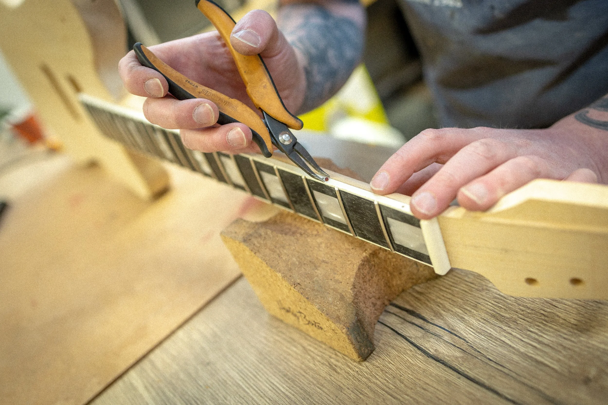 Handen snijdt een stuk van een gitaarhanger met een pincet, op een werkbank met hout.