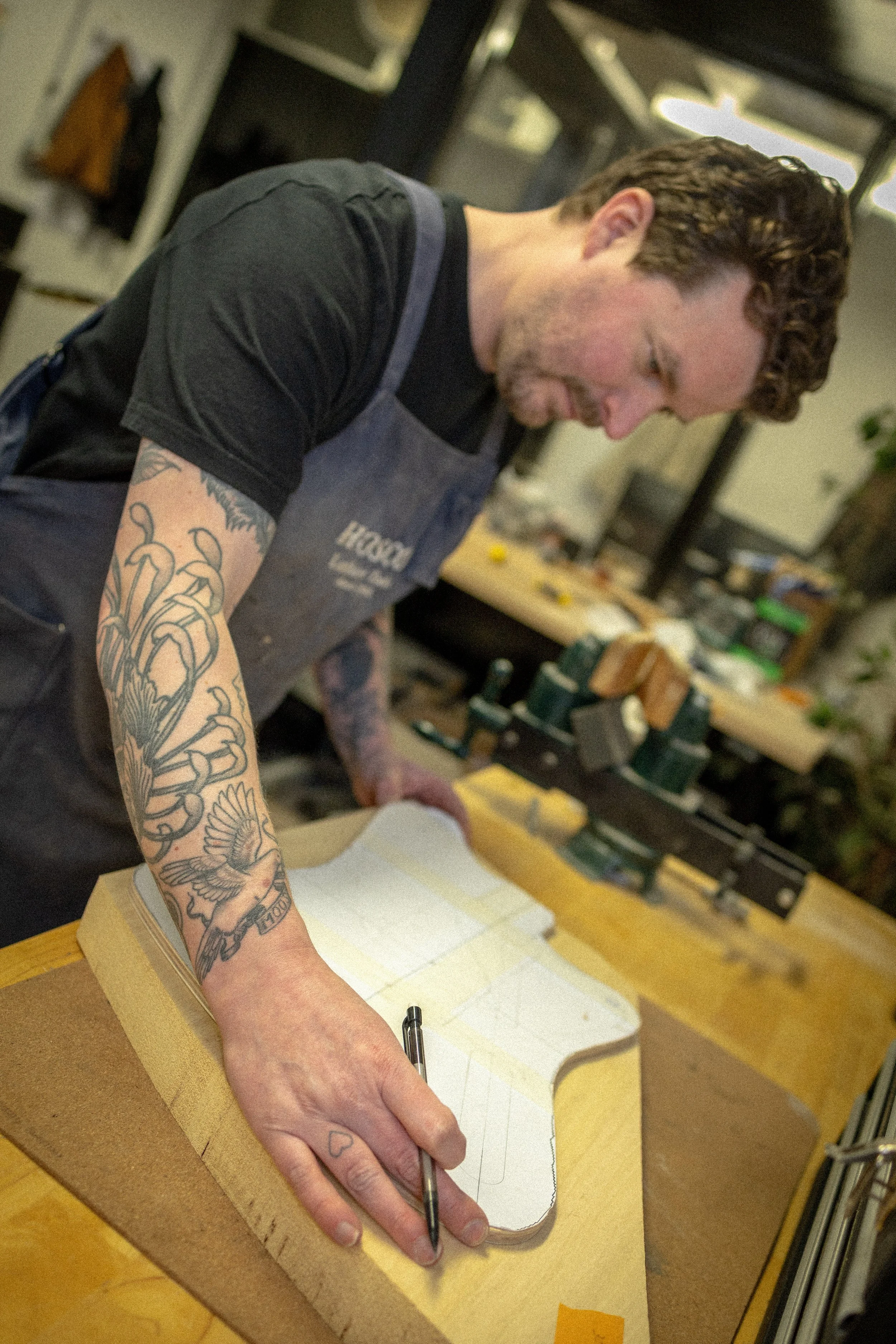 Een persoon met getatoeëerde armen die een potlood vasthoudt en bezig is met het tekenen op een stuk papier dat op een stevige ondergrond ligt. De achtergrond toont een werkruimte met gereedschap en dozen.