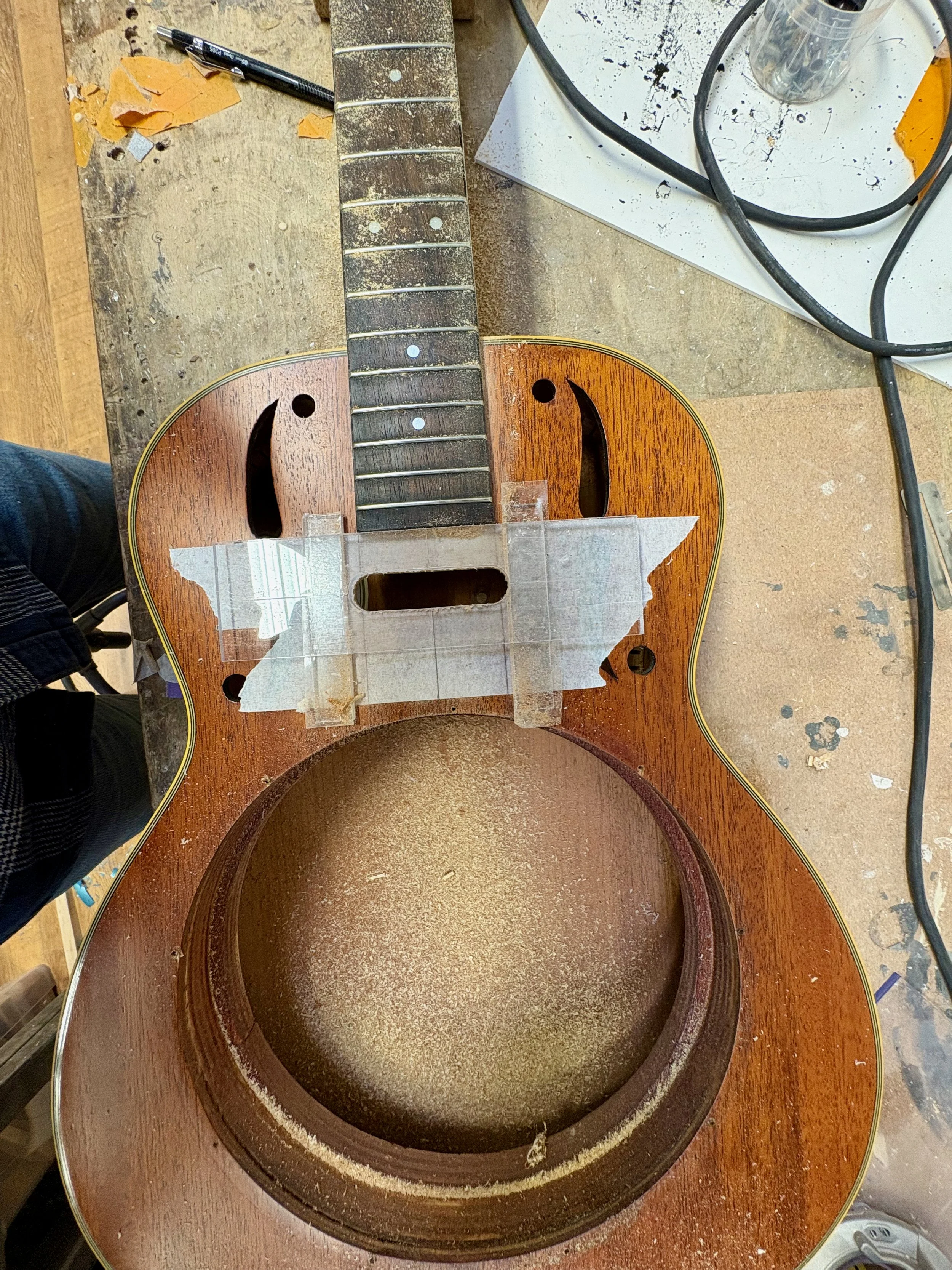 Een houten werkbank met een onafgewerkte gitaar, omringd door gereedschap en papieren. Er ligt tape en een scherp mes op de tafel.