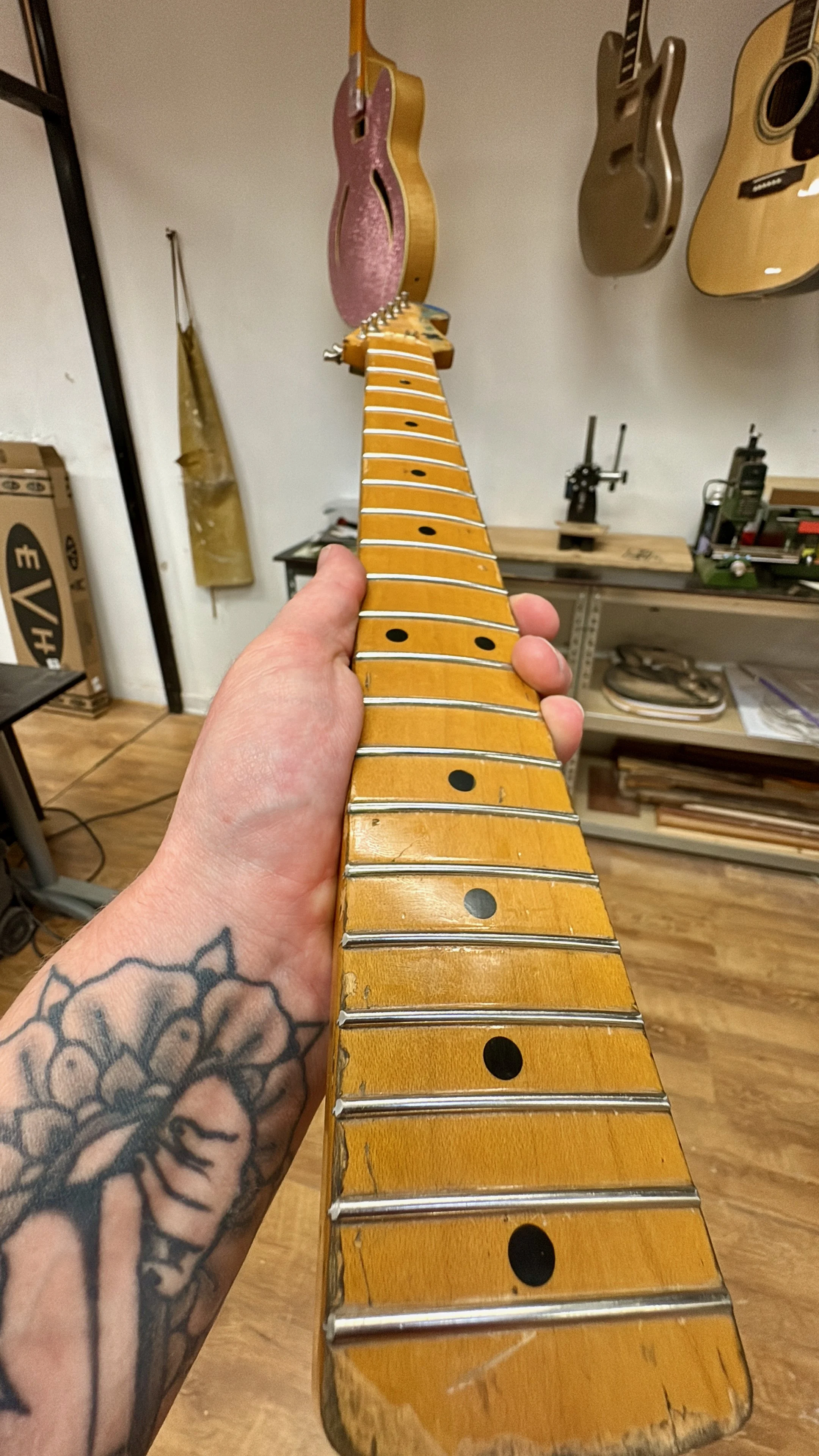 Close-up van een gitaarhals met blanke snaren en vingerkuipen, terwijl iemand met een tattoo op de arm de gitaar vasthoudt.