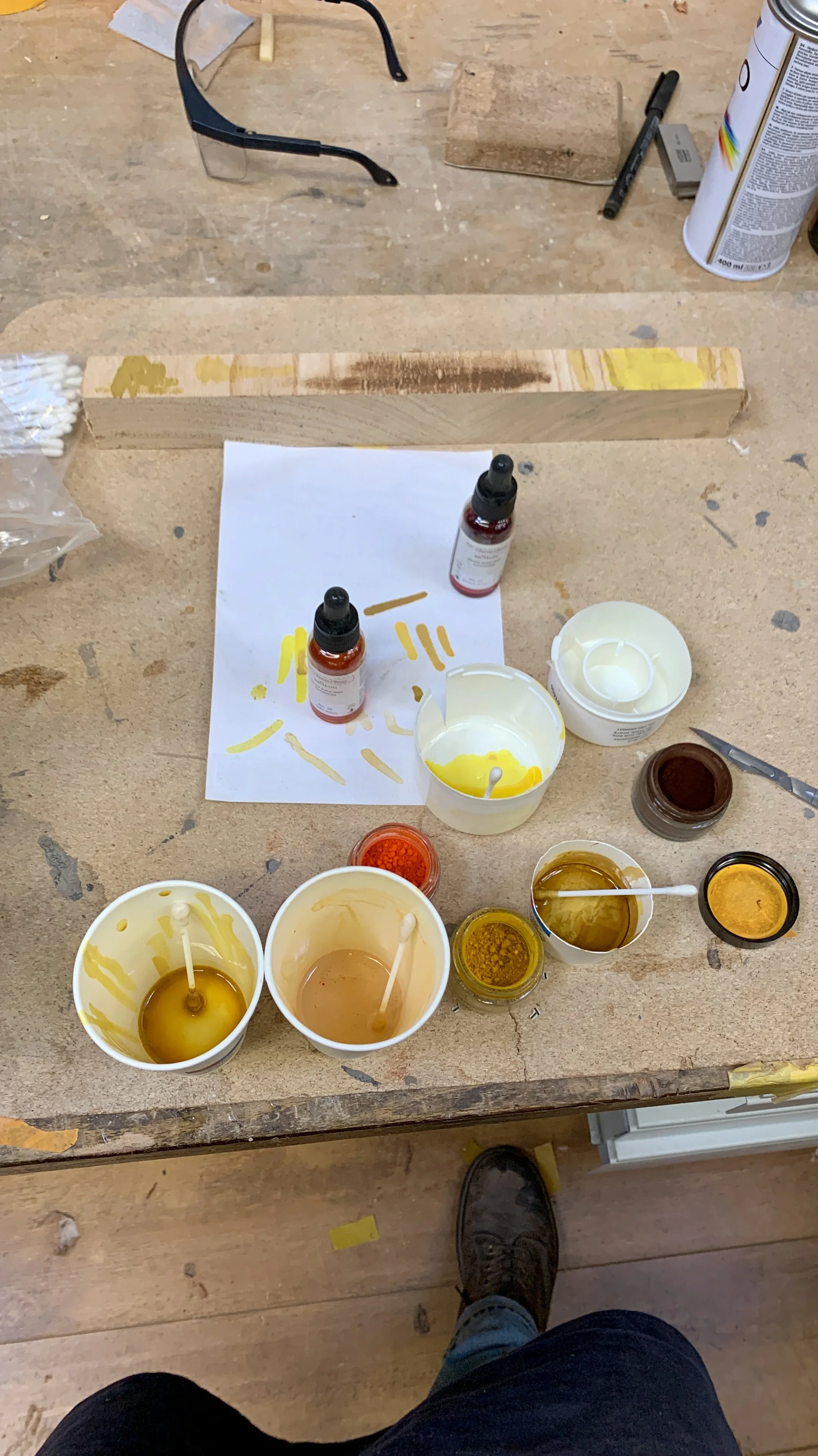 Wereld van verf en kunstbenodigdheden op een werkbank, inclusief potten verf, lepels en verfbollen, met een persoon die de foto neemt en een voet zichtbaar.