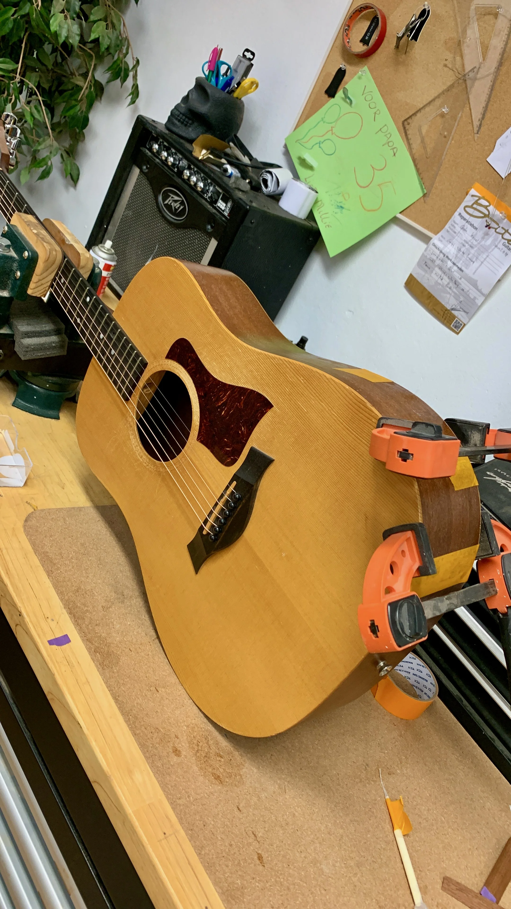 Gitaartje op een werkbank met verschillende gereedschappen en materiaal rondom, waaronder houtklemmen en een kladblok met aantekeningen.