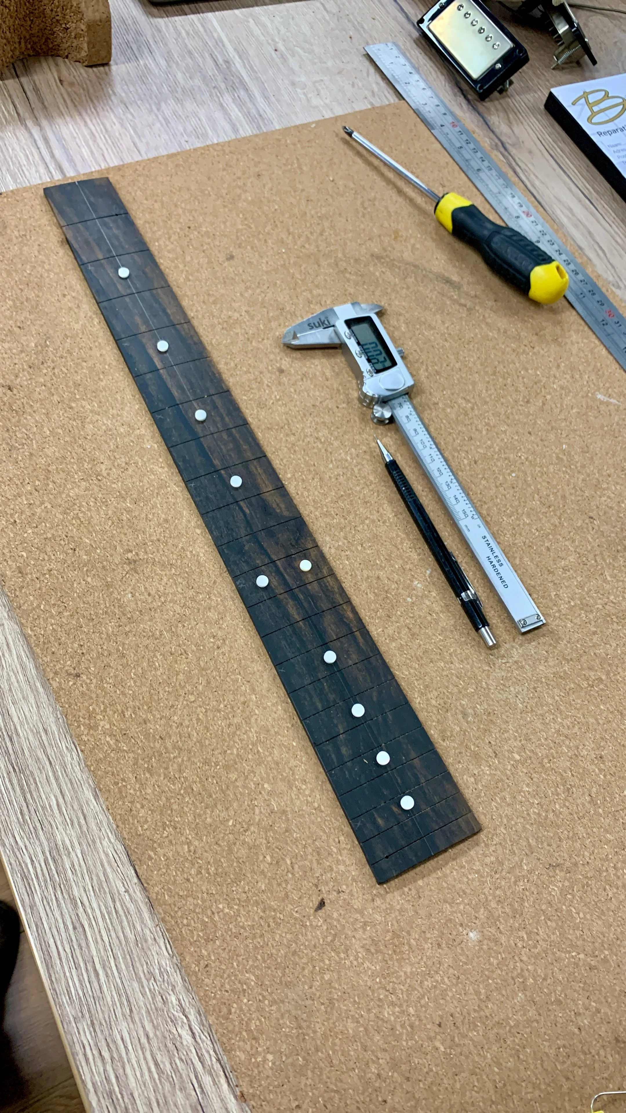 Werkblad met meetinstrumenten, waaronder een digitale schuifmaat, een liniaal, een schroevendraaier en een stuk hout. Op de achtergrond liggen onderdelen voor een elektronisch apparaat.