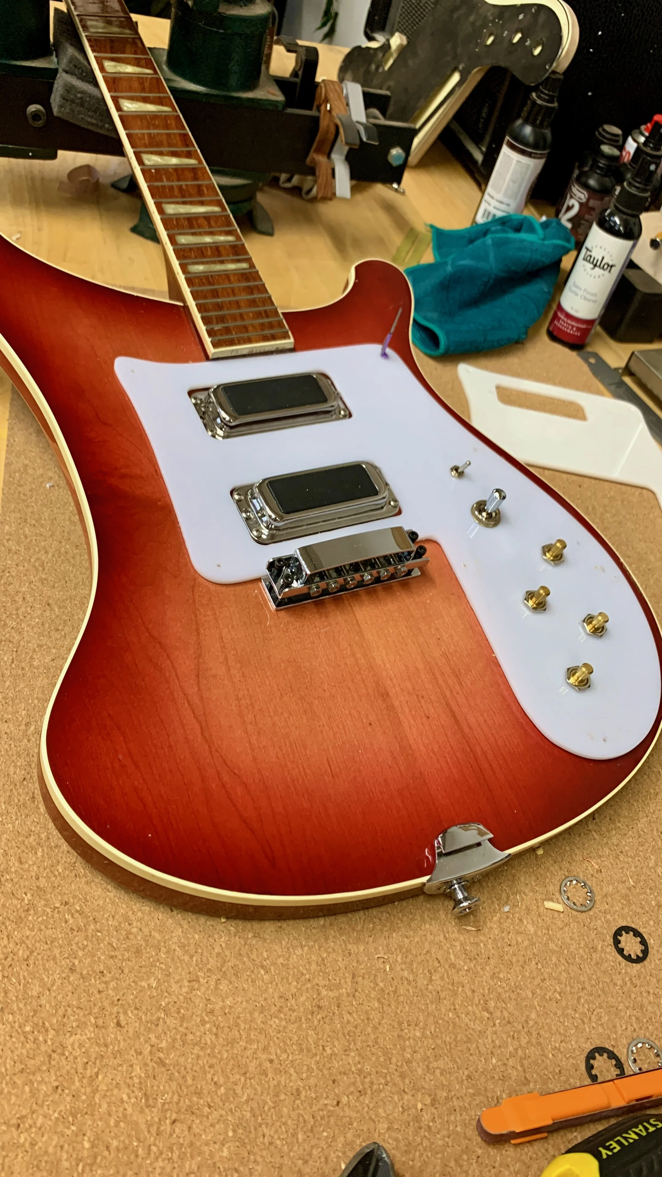 Een rode elektrische gitaar zonder snaren, met een witte pickguard en elektrische componenten, op een werkbank met gereedschap en andere onderdelen.