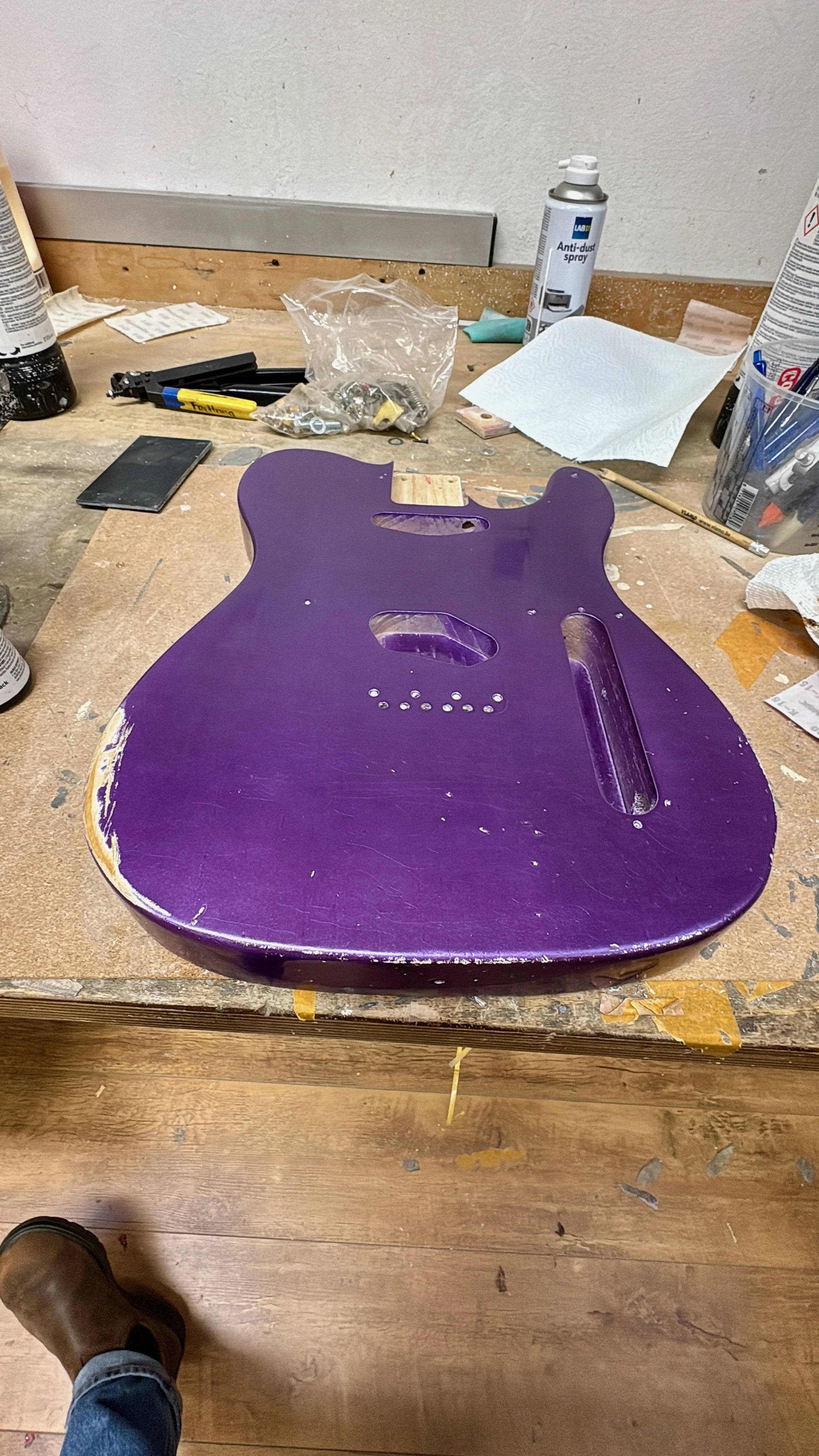 Een werktafel met een ongeassembleerde elektrische gitaar body in paars, omringd door gereedschap, stoffen en verfblekken.