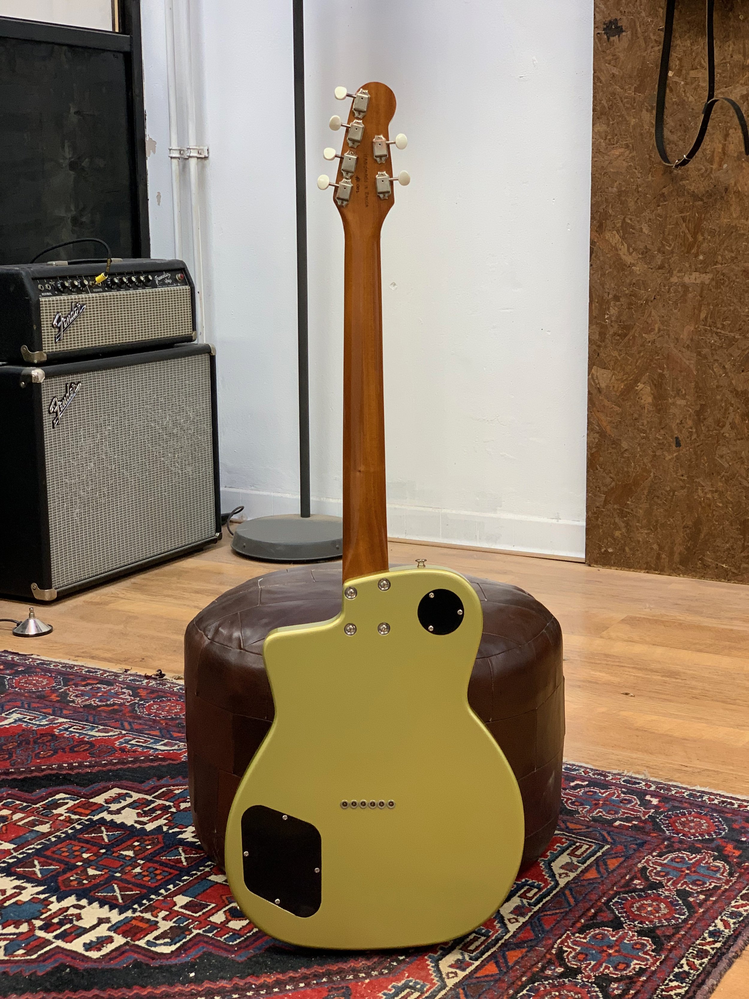 Elektrische gitaar met gele body en houten hals, geplaatst op een ronde, bruin, gestikte zitkussen. Op de achtergrond staan een fender gitaarversterker en een gitaarstandaard. De ruimte heeft houten vloer en een paar nuance wanden met een tapijt op d