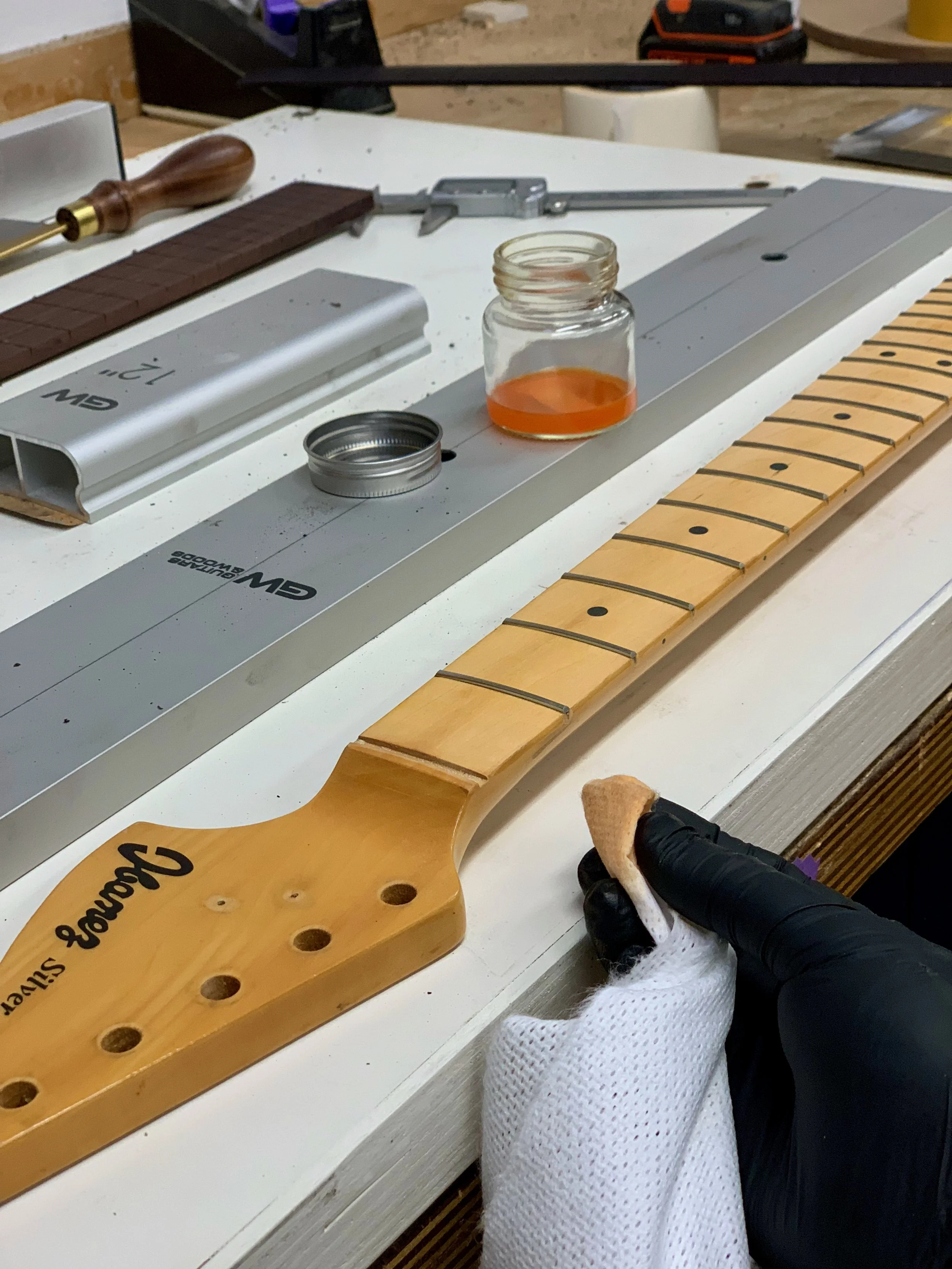 Een persoon die een gitaar afstelt, houdt een doek of borstel vast om het fretboard schoon te maken. Op de werkbank liggen gereedschap en onderdelen van een gitaar, inclusief een hals met frets en een open potje met oranje lak of olie.