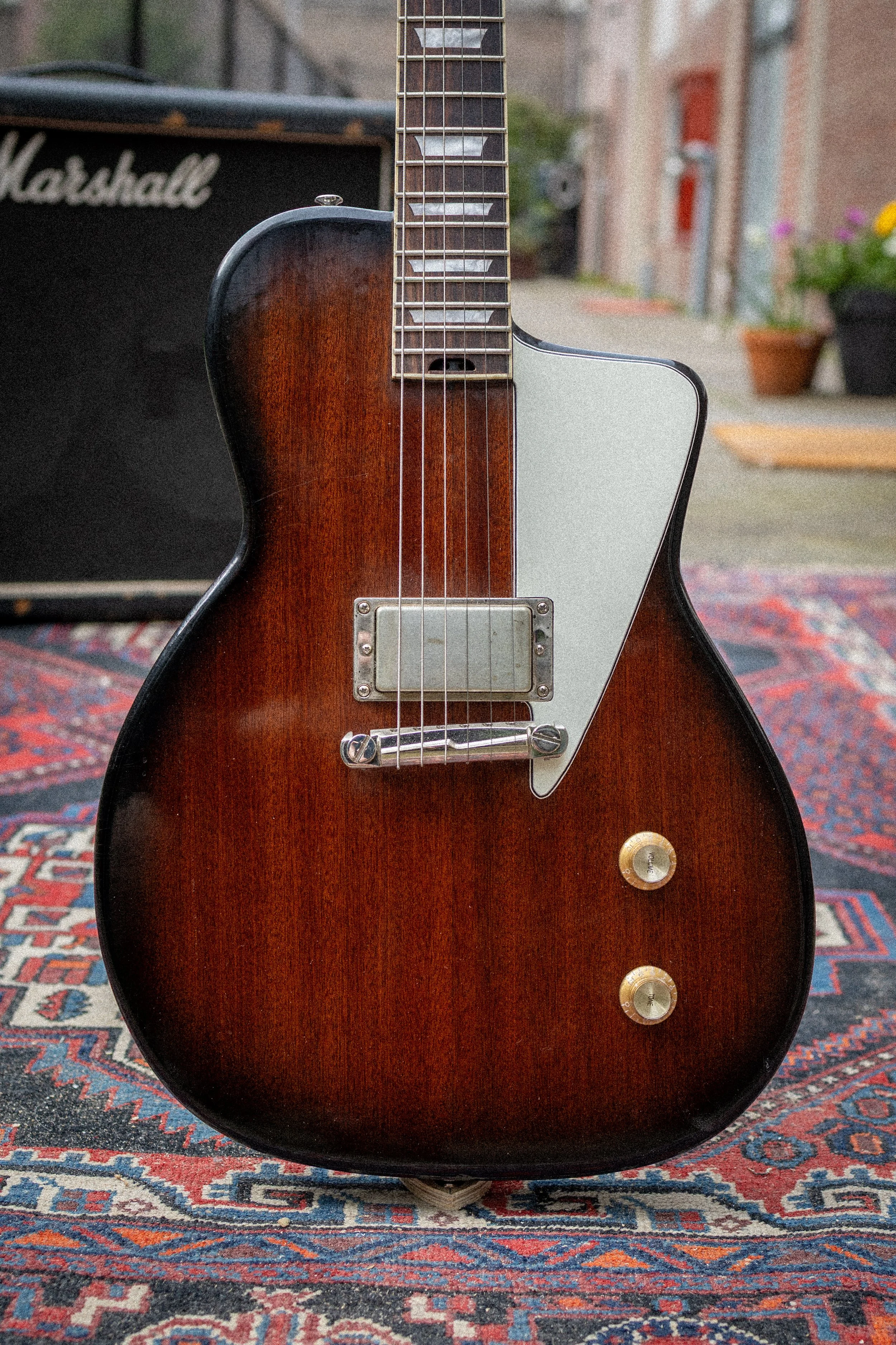 Foto van een elektrische basgitaar met een bruine vlam en een witte slagplaat, staand op een tapijt met een gitaarversterker op de achtergrond in een buitenomgeving.