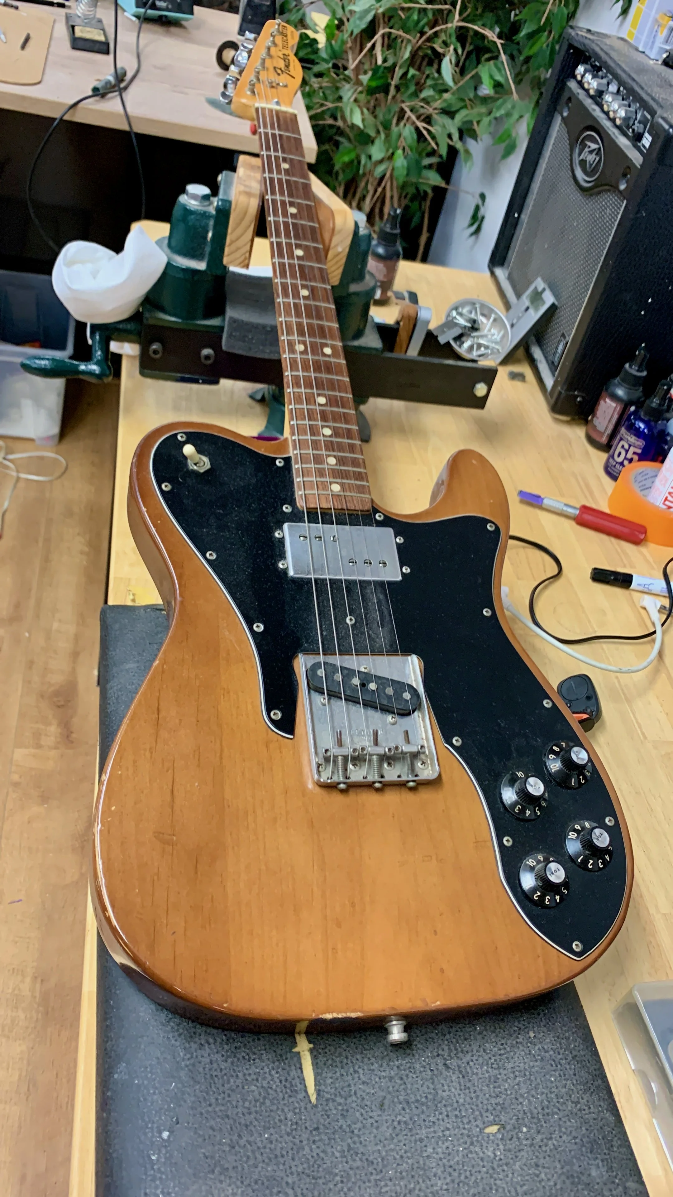 Een elektrische gitaar met een houten afwerking ligt op een werkbank in een werkplaats, omgeven door gereedschap en apparatuur.