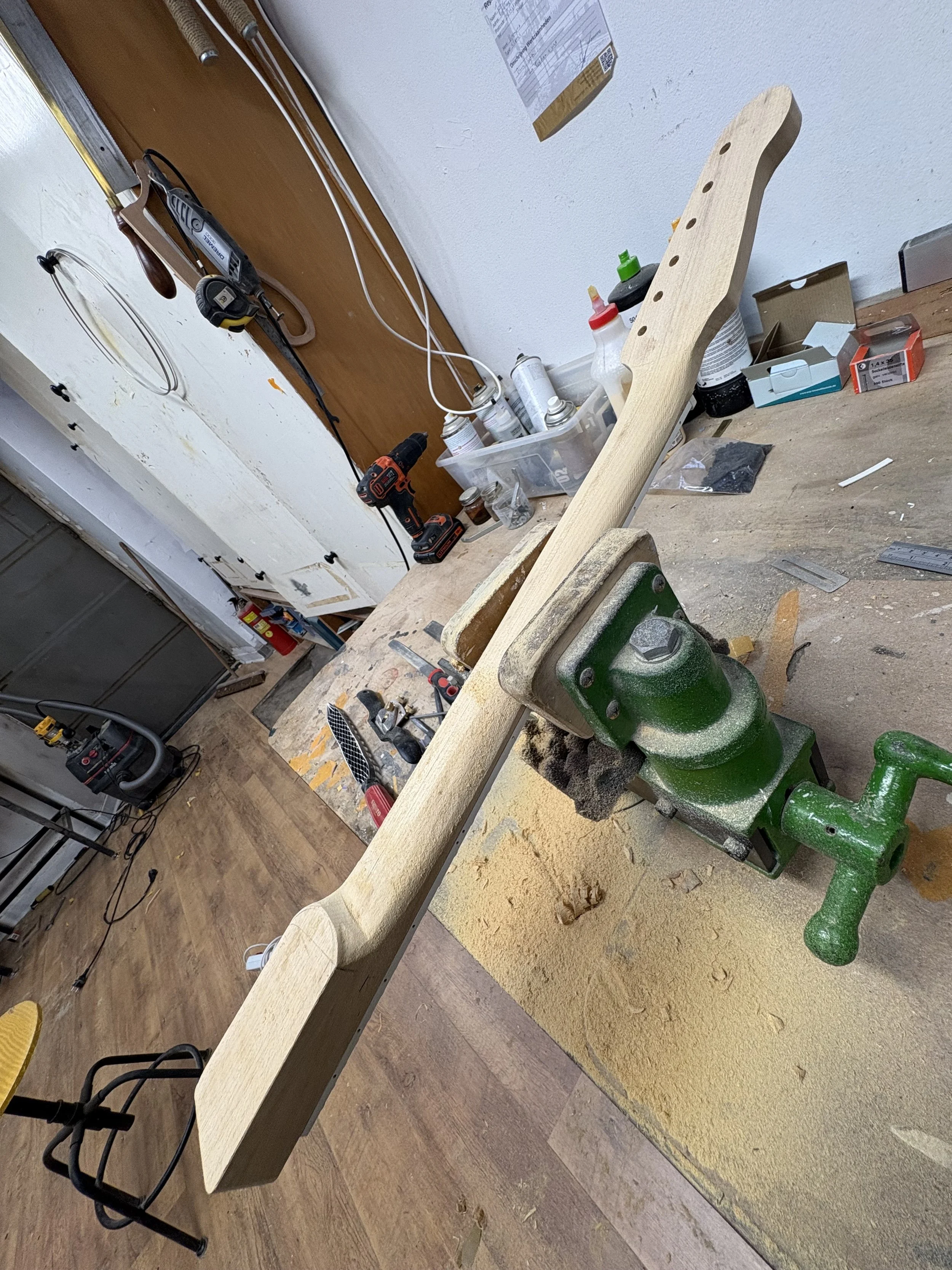 Een werkbank met een houten stuk dat vastgezet is in een groene bankschroef, diverse gereedschappen, verfspuiten, zand en verpakkingen op en rond de werkbank.